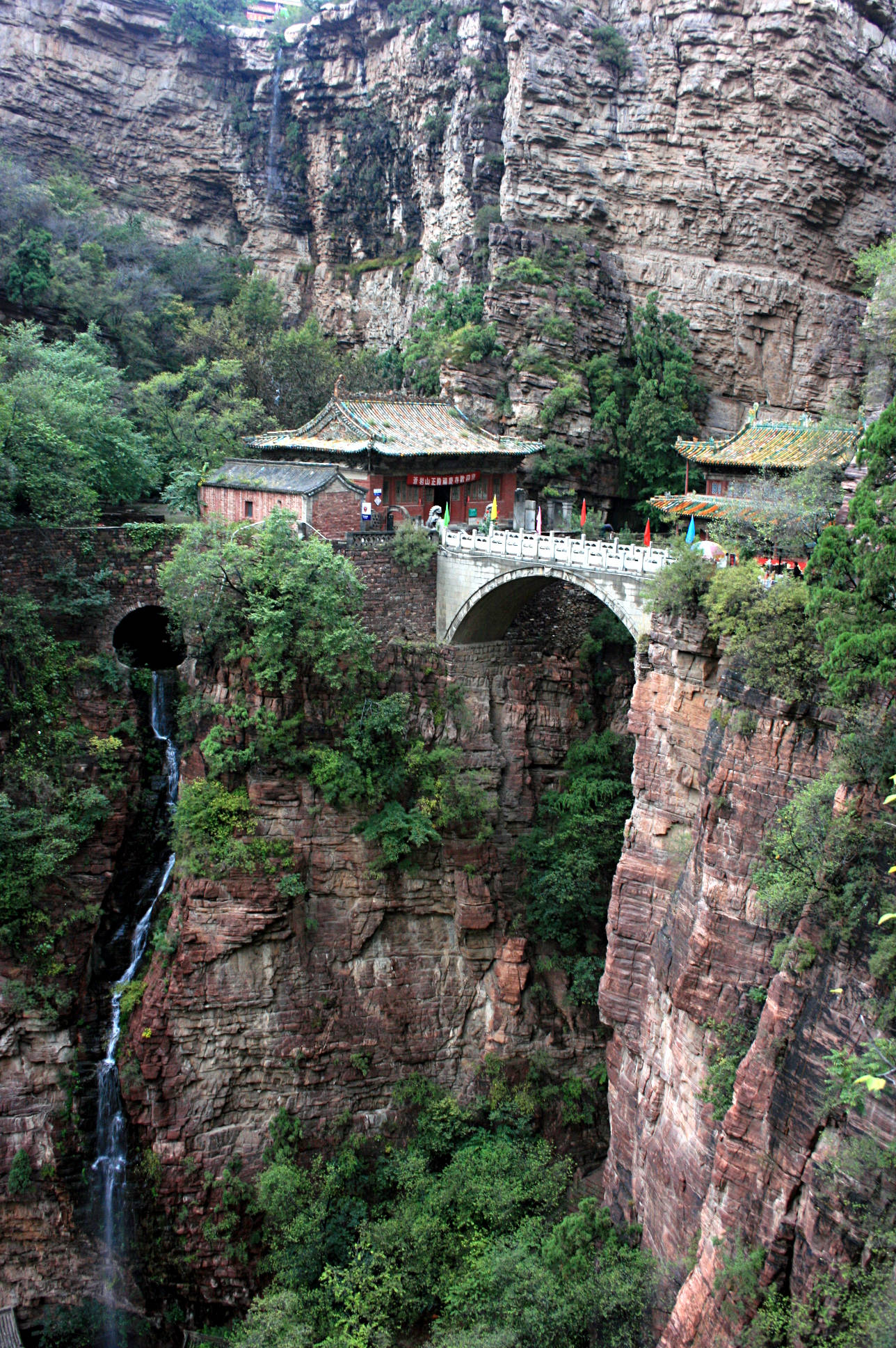 中國 桌布 壁紙 — 一座傳統中國寺廟坐落在懸崖邊，石橋拱跨瀑布和茂密綠植。