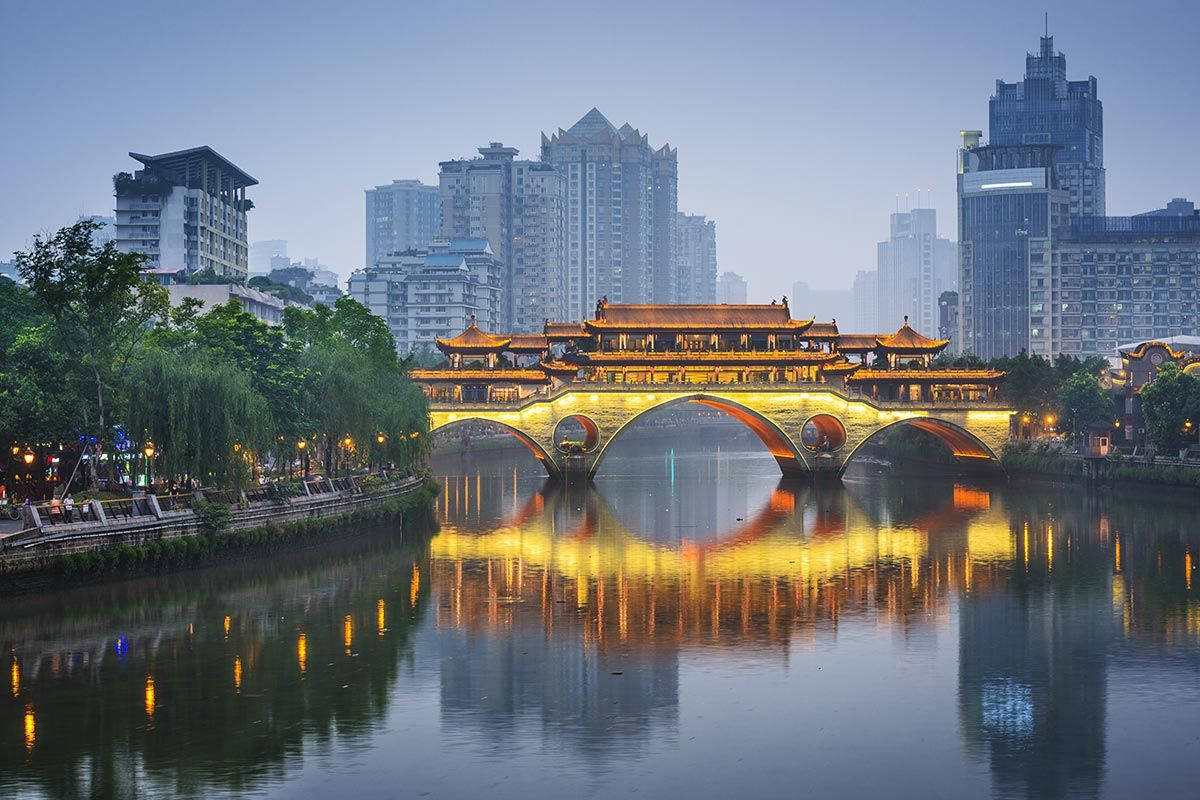 Chengdu 桌布 壁紙 — 一座傳統中國橋樑與發光的燈籠在平靜的水面上美麗地倒映，天空暮色被現代摩天大樓框住。
