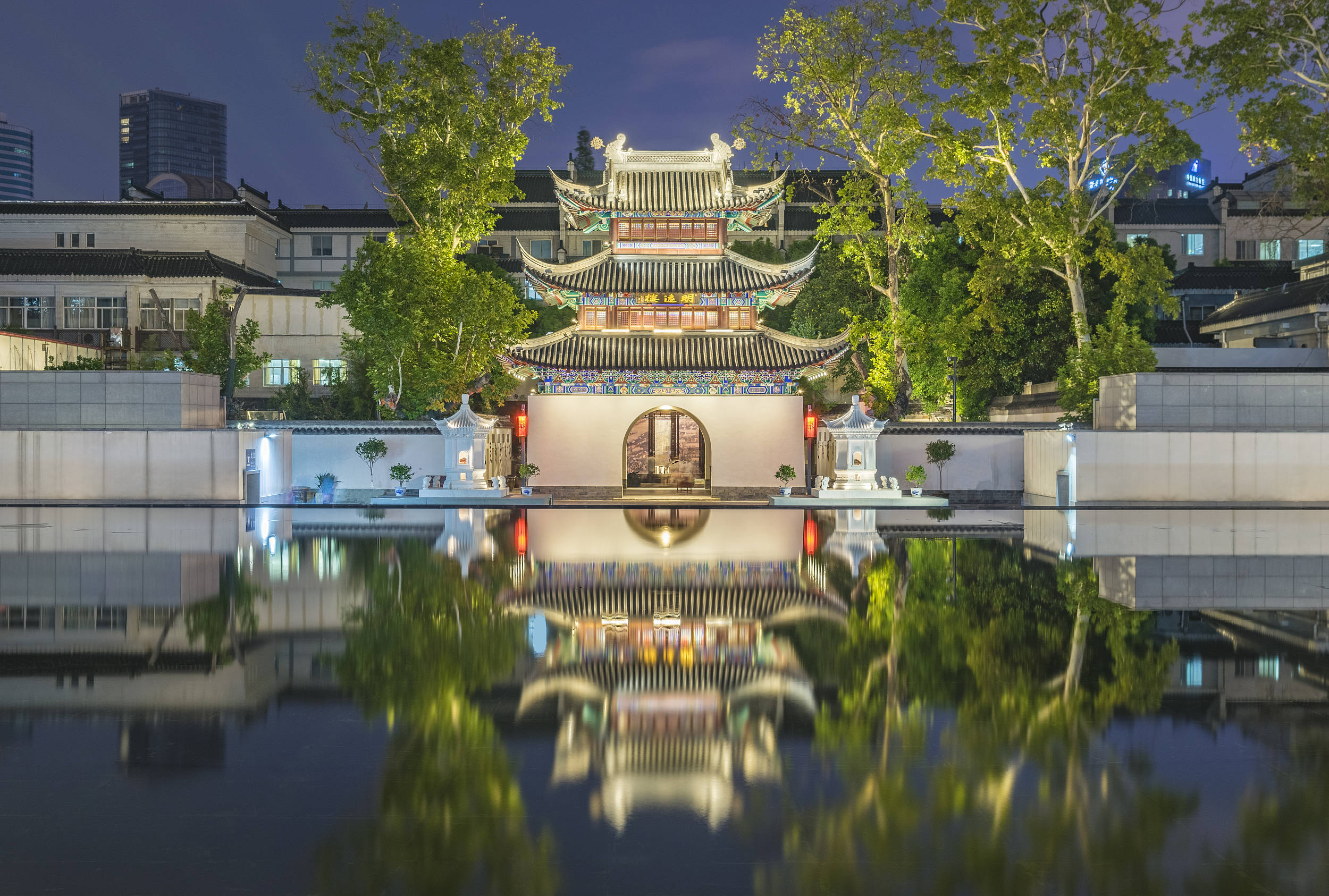 Nanjing 桌布 壁紙 — 一座傳統中式亭台樓閣，飛簷翹角，在夜晚被美麗地照亮，其倒影完美地映照在平靜的水池中，四周環繞著鬱鬱蔥蔥的綠樹。
