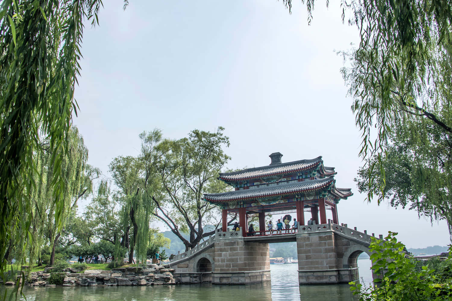 樹木 桌布 壁紙 — 一座傳統中式亭橋拱跨在平靜的水面上，被垂柳枝條和茂密的綠樹環繞，映襯著明亮的天空。