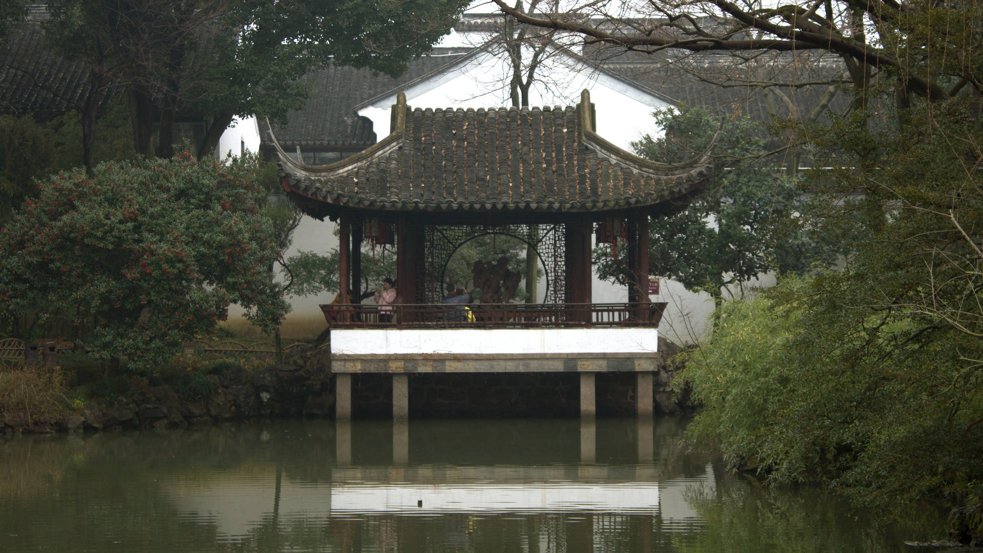 Suzhou 桌布 壁紙 — 一座傳統中式亭閣，擁有彎曲屋頂，立於平靜水面的支柱上，四周環繞著茂密的綠植和光禿的樹木。