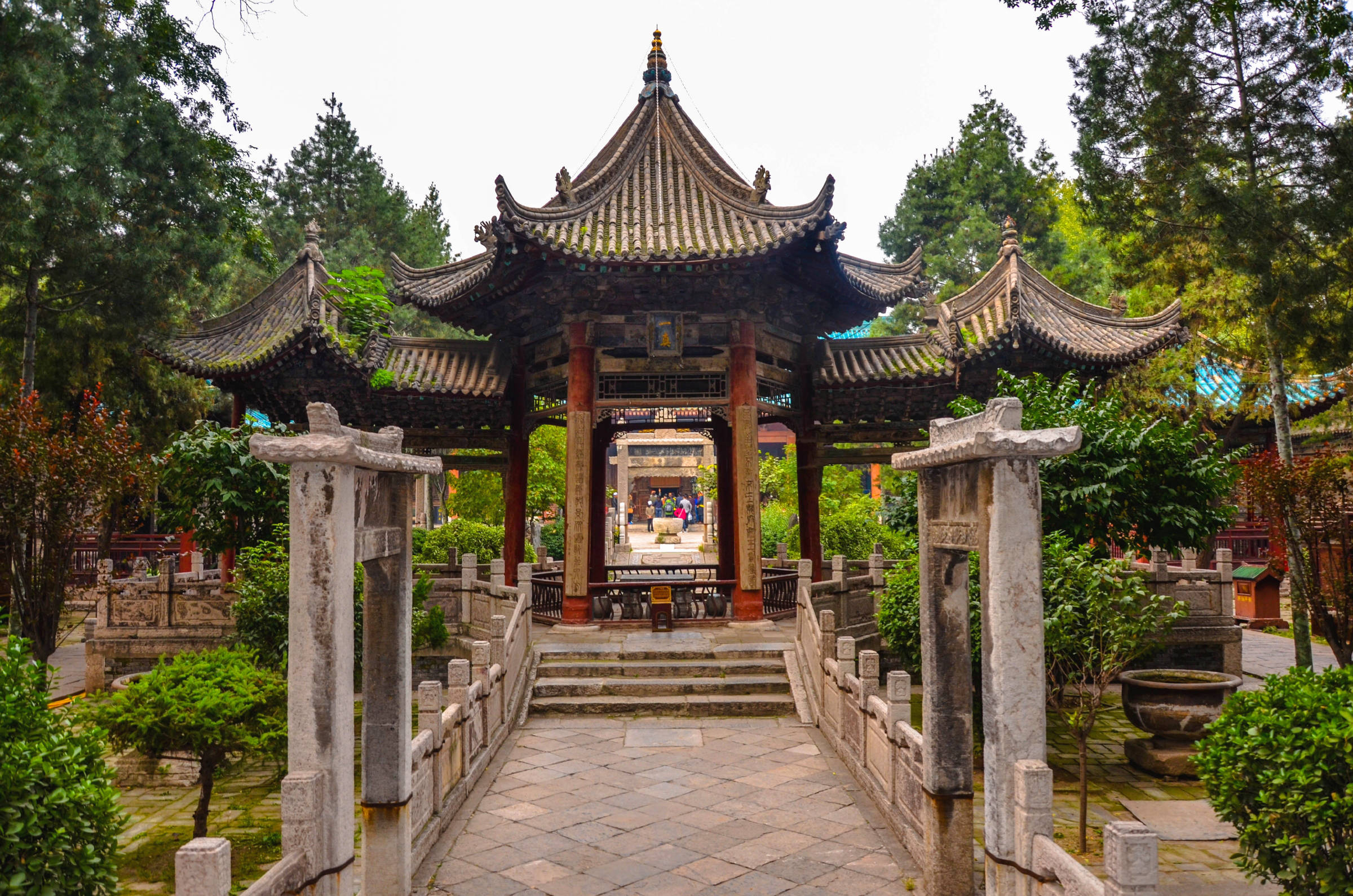 Xian 桌布 壁紙 — 一座傳統中式亭閣，擁有彎曲的瓦片屋頂，矗立在石橋盡頭，周圍環繞著茂密的綠樹和灌木。