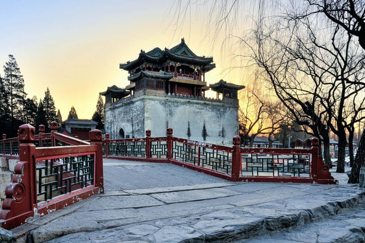 建築 桌布 壁紙 — 一座傳統中式亭閣，綠色屋頂和紅色欄杆，在日落時分立於石橋後方，周圍是光禿的樹木和柔和的金色光芒。