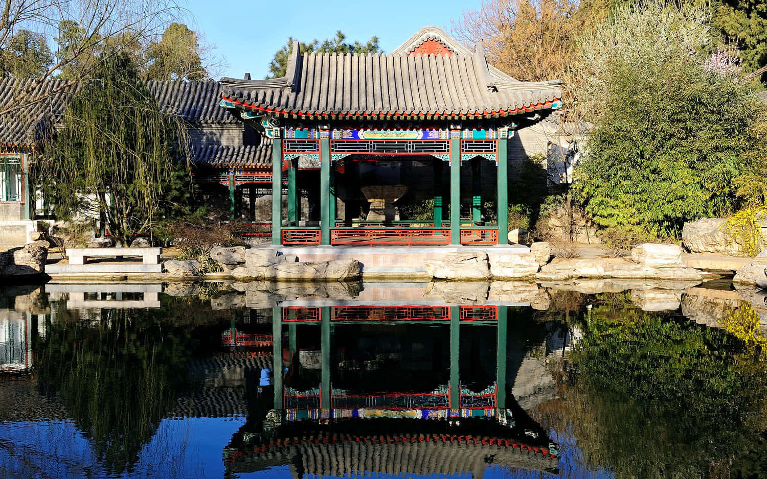 建築 桌布 壁紙 — 一座傳統中式亭閣，綠色柱子配紅色點綴，佇立於平靜池塘旁，在清澈藍天下倒映出其精緻細節。
