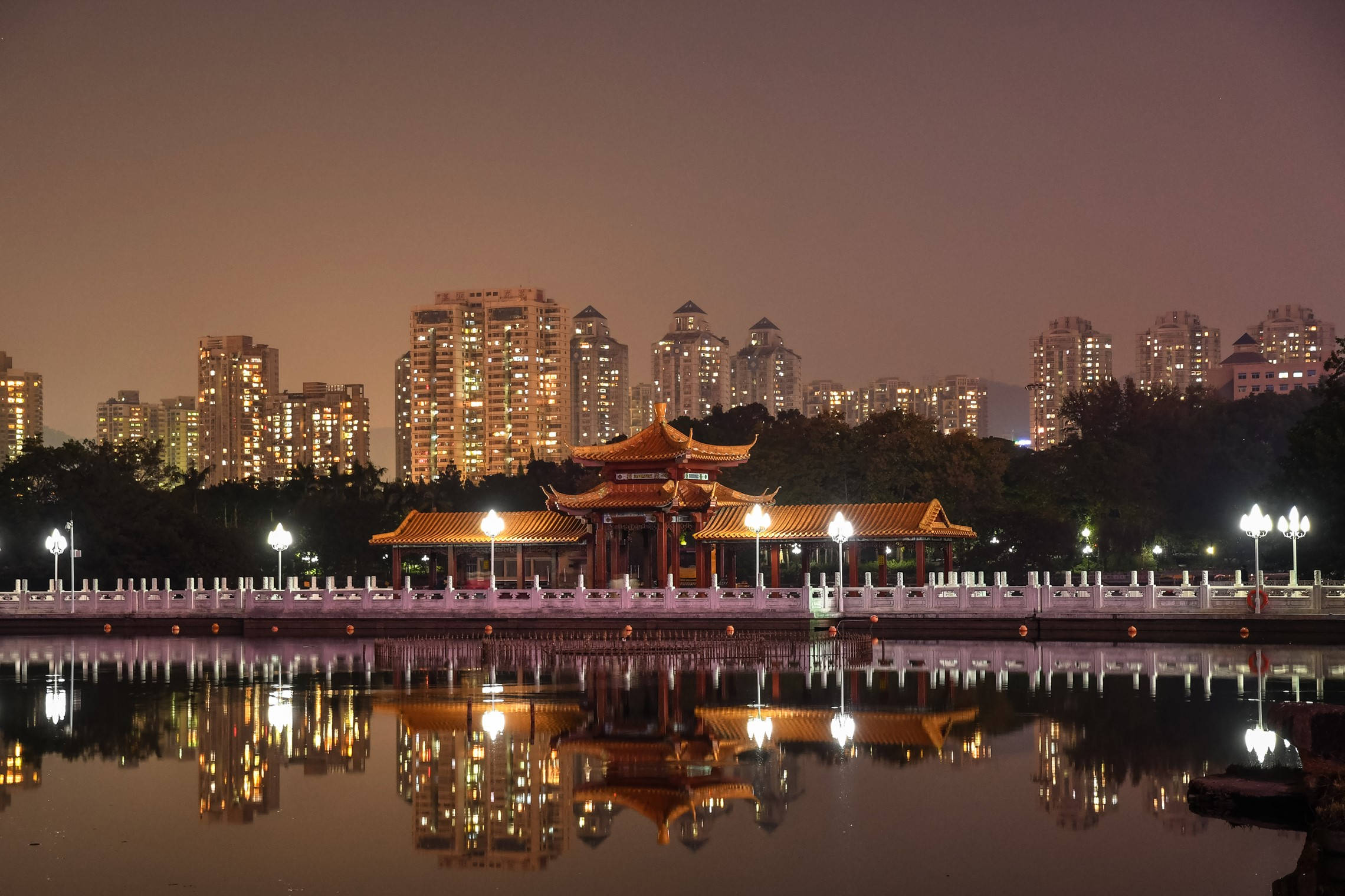 Shenzhen 桌布 壁紙 — 一座傳統中式亭閣在夜晚溫暖地發光，其倒影完美地映照在平靜的水面上，背景是燈火通明的城市摩天大樓。