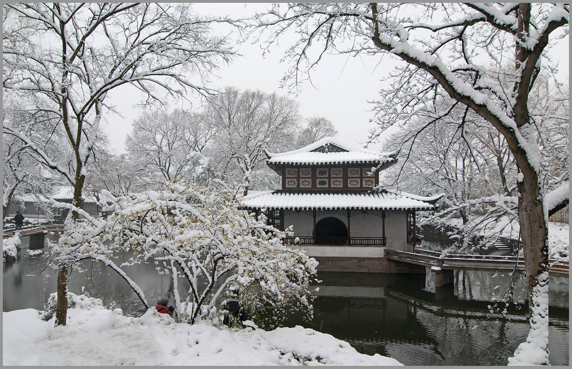 Suzhou 桌布 壁紙 — 一座傳統中式亭閣在雪中靜立，被雪壓枝頭的樹木環繞，旁邊是一池平靜的湖水。