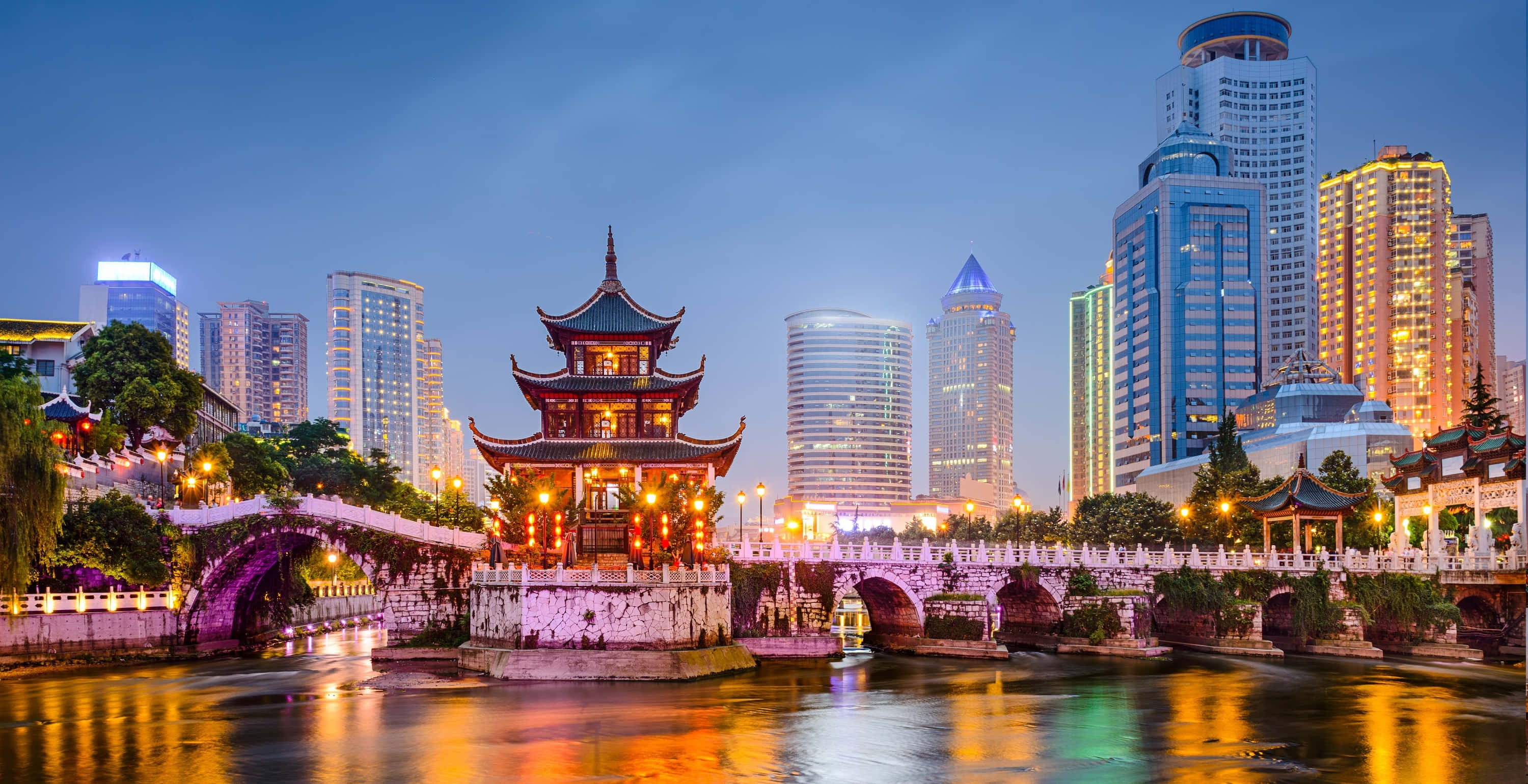 中國 桌布 壁紙 — 一座傳統中式亭閣在黃昏的河上溫暖地發光，被水中倒映的現代摩天大樓所環繞。