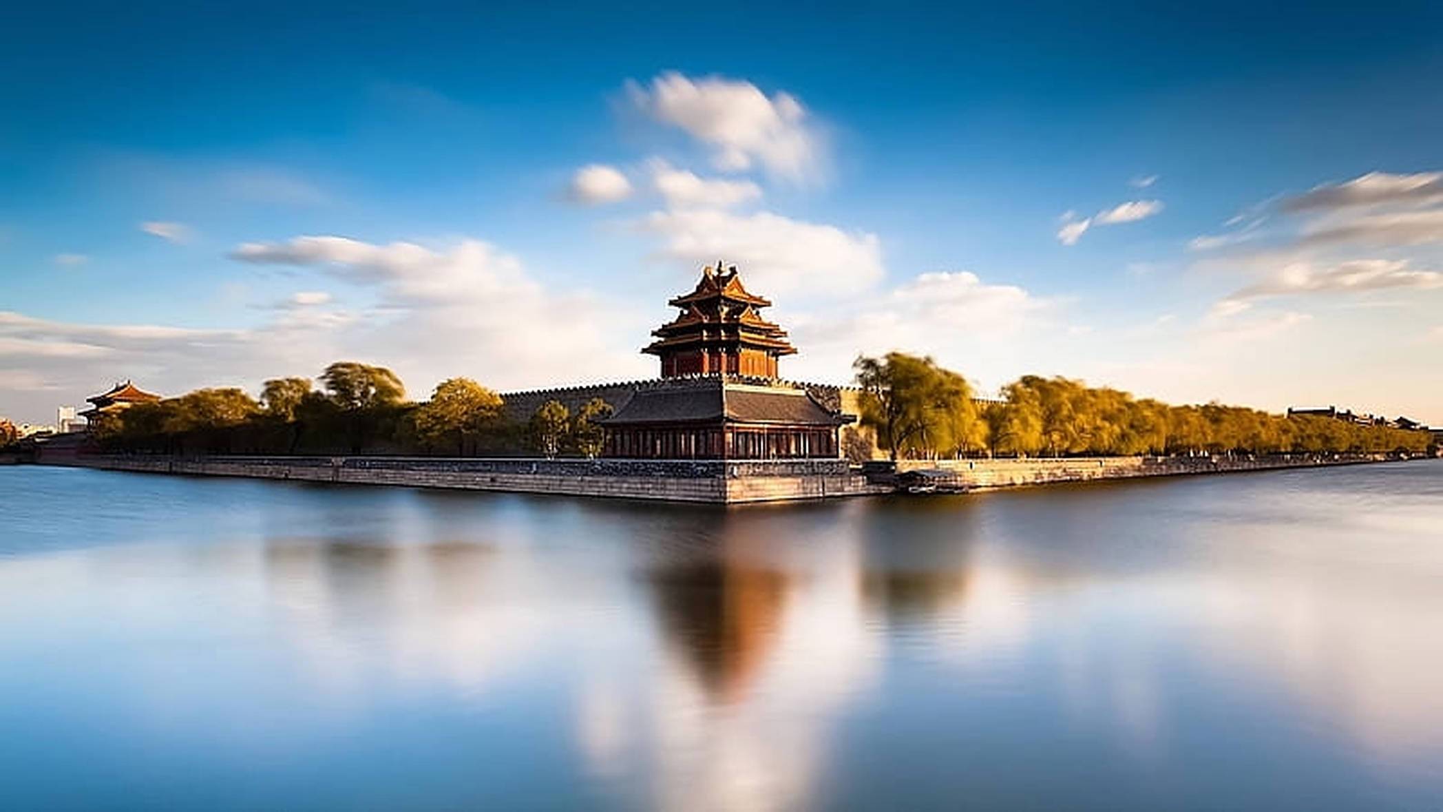 Wuhan 桌布 壁紙 — 一座傳統中式宮殿塔樓巍然聳立於石砌平台上，四周環繞平靜水面，上方是湛藍天空與零星白雲。