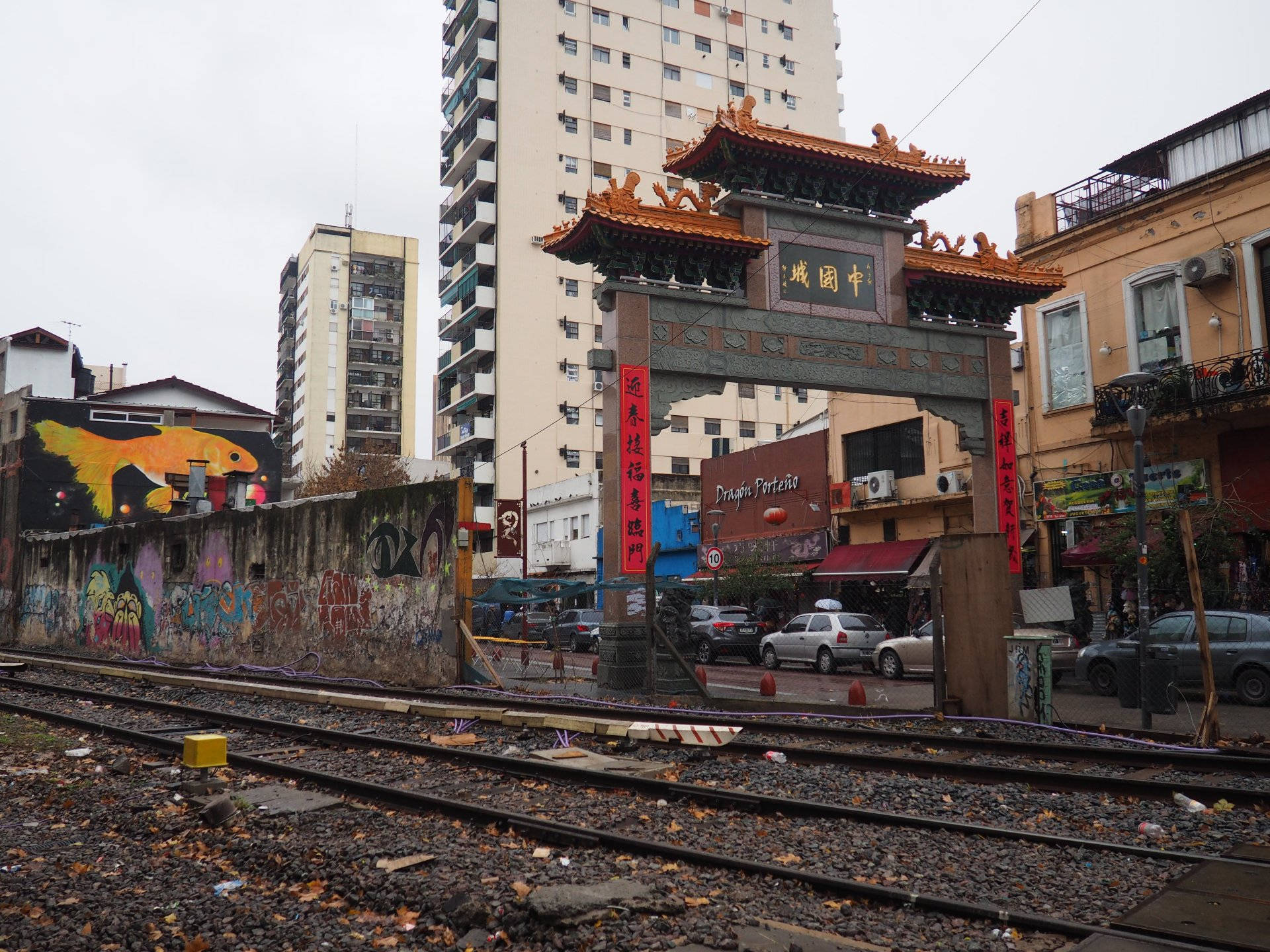 Aires 桌布 壁紙 — 一座傳統中式牌坊矗立於城市衰敗之中，前景是塗鴉牆壁和火車軌道，天空陰沉。