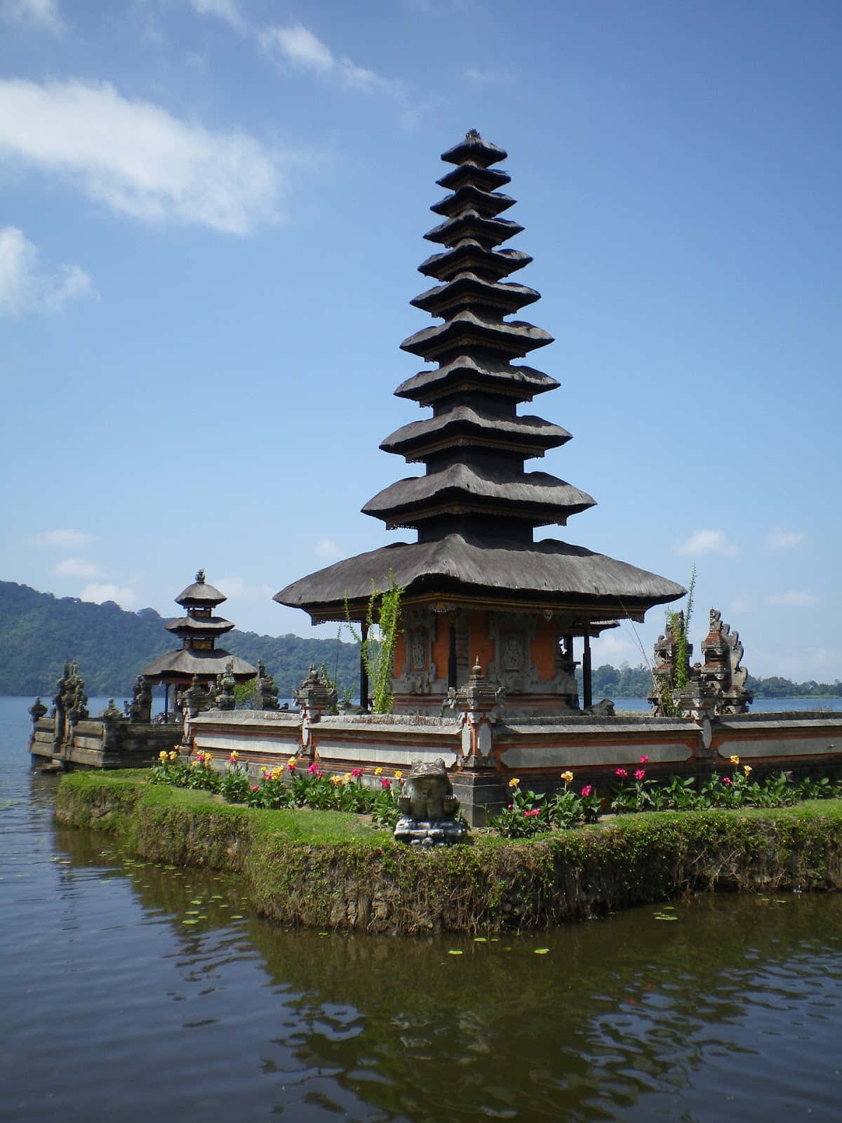 Bali 桌布 壁紙 — 一座傳統峇里島寺廟，擁有層疊屋頂，寧靜地矗立於充滿水的島嶼上，周圍環繞著茂密的綠植和鮮豔的花朵，背景是清澈的藍天。