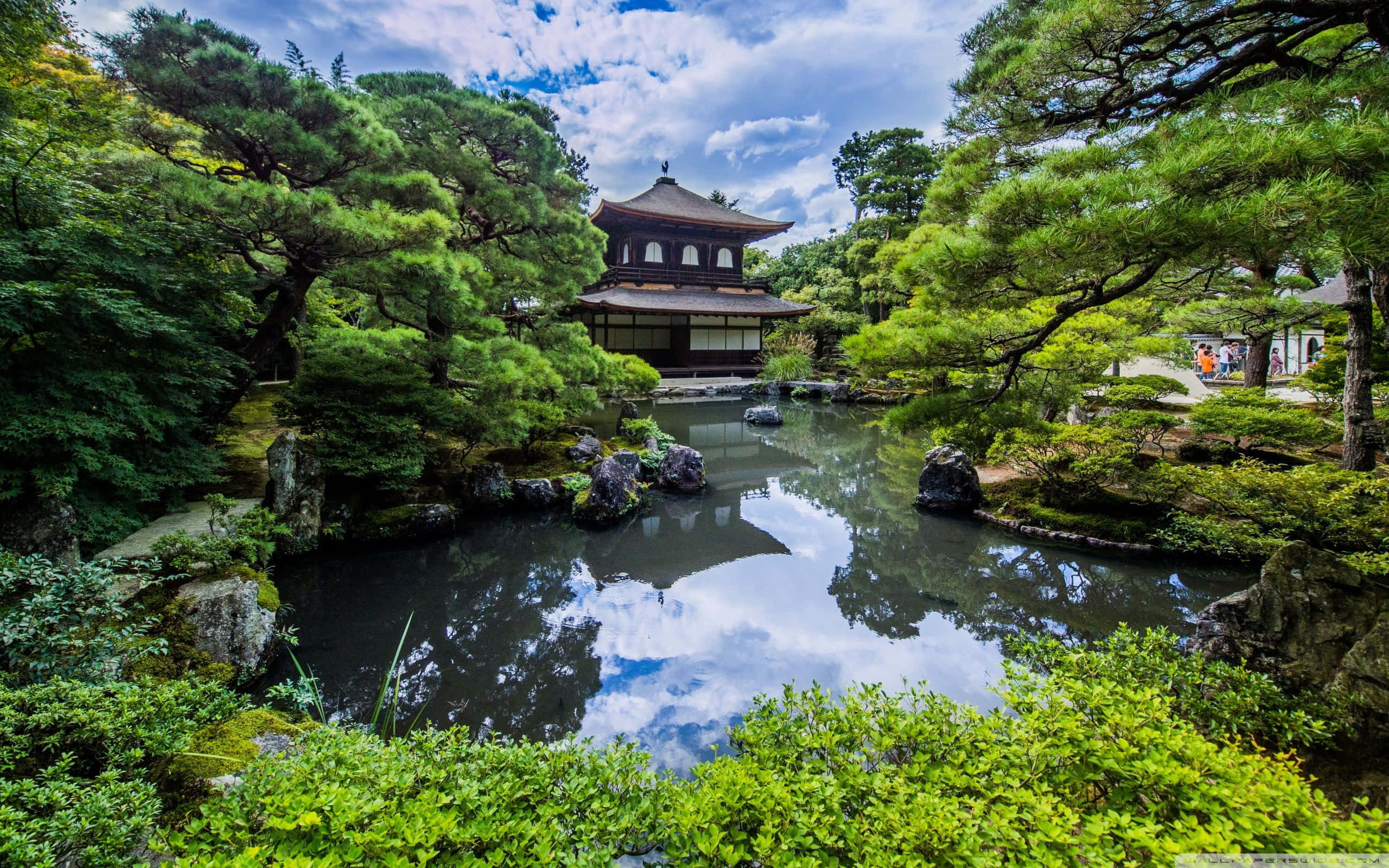 Japan Landscape 桌布 壁紙 — 一座傳統日本亭閣倒映在寧靜的池塘中，周圍環繞著茂密的綠樹和長滿苔蘚的岩石，天空部分多雲。