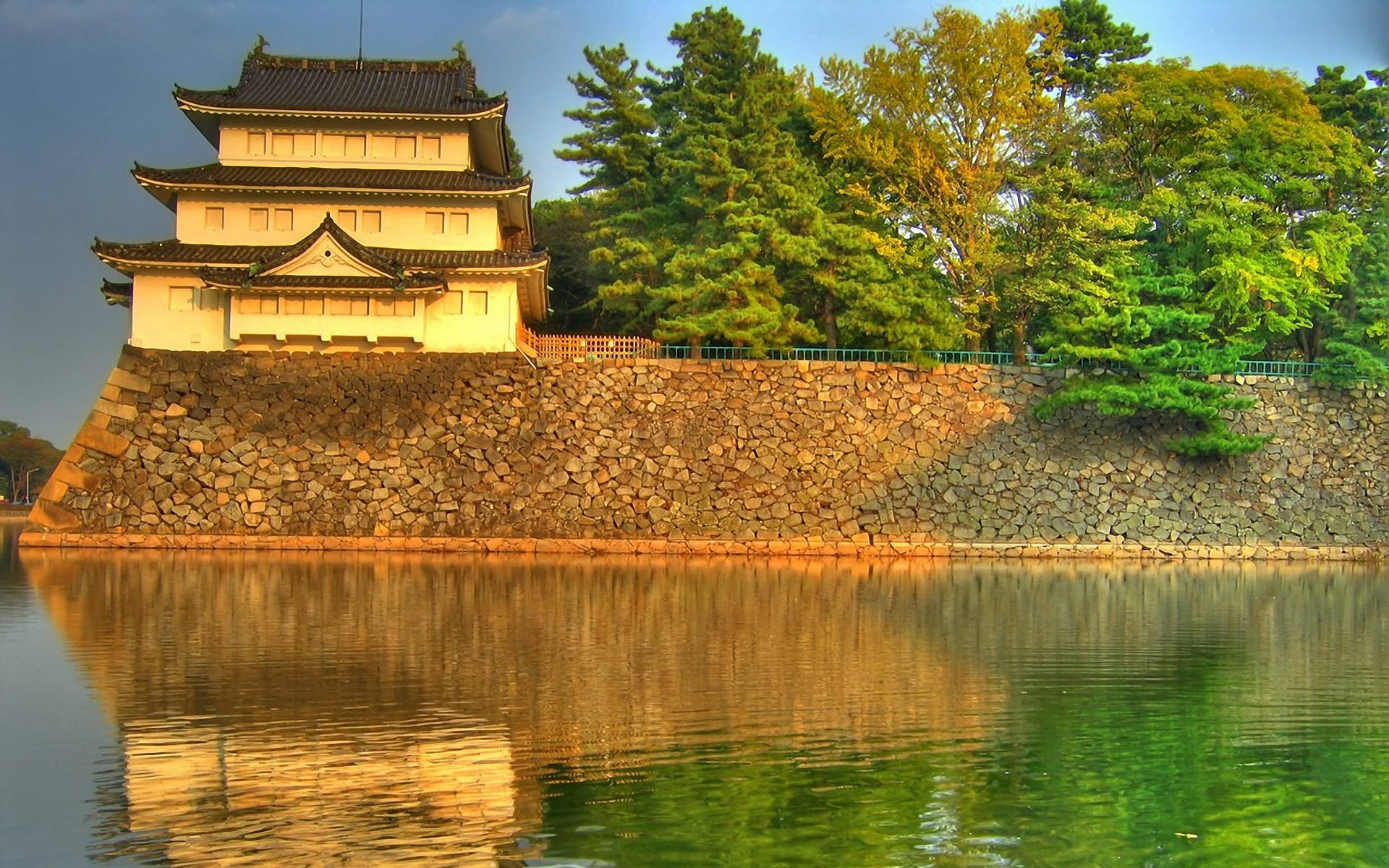 中式房屋壁紙 桌布 壁紙 — 一座傳統日本城堡，擁有層疊屋頂，矗立在石牆上，旁邊平靜的水面倒映著金色光芒和茂密的綠樹。