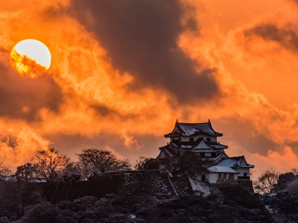 Himeji Castle 桌布 壁紙 — 一座傳統日本城堡在火紅橙色夕陽天空的襯托下呈現剪影，一輪發光的太陽部分被雲層遮擋。