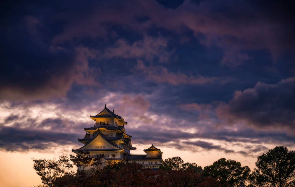 Himeji Castle 桌布 壁紙 — 一座傳統日本城堡在紫色和橙色雲彩的戲劇性暮色天空下被照亮。
