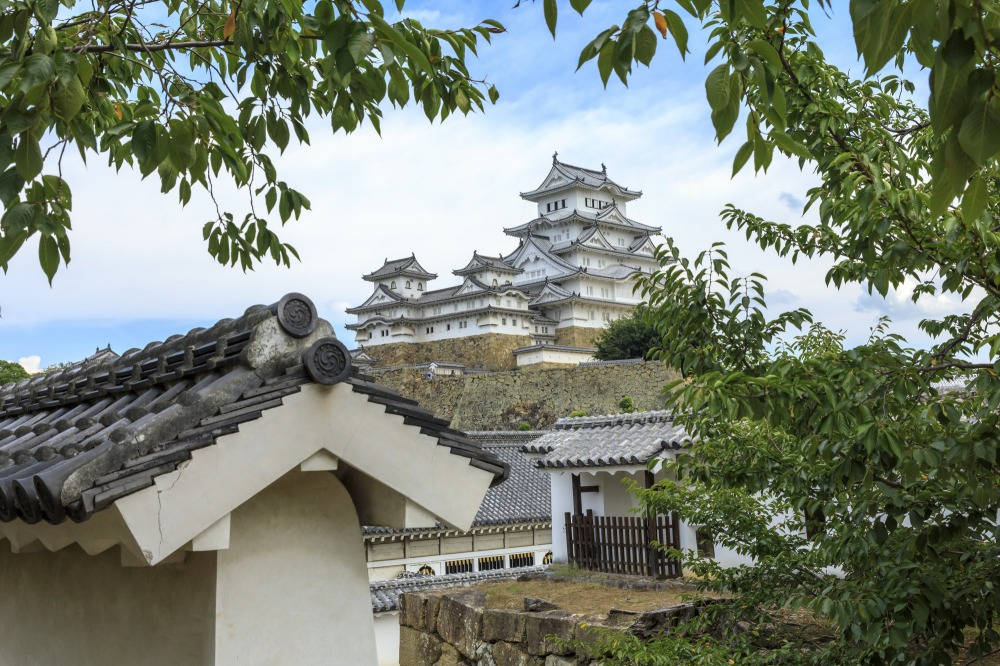 Himeji Castle 桌布 壁紙 — 一座傳統日本城堡在茂密綠葉和瓦片屋頂後方，於明亮的藍天下雄偉地聳立。