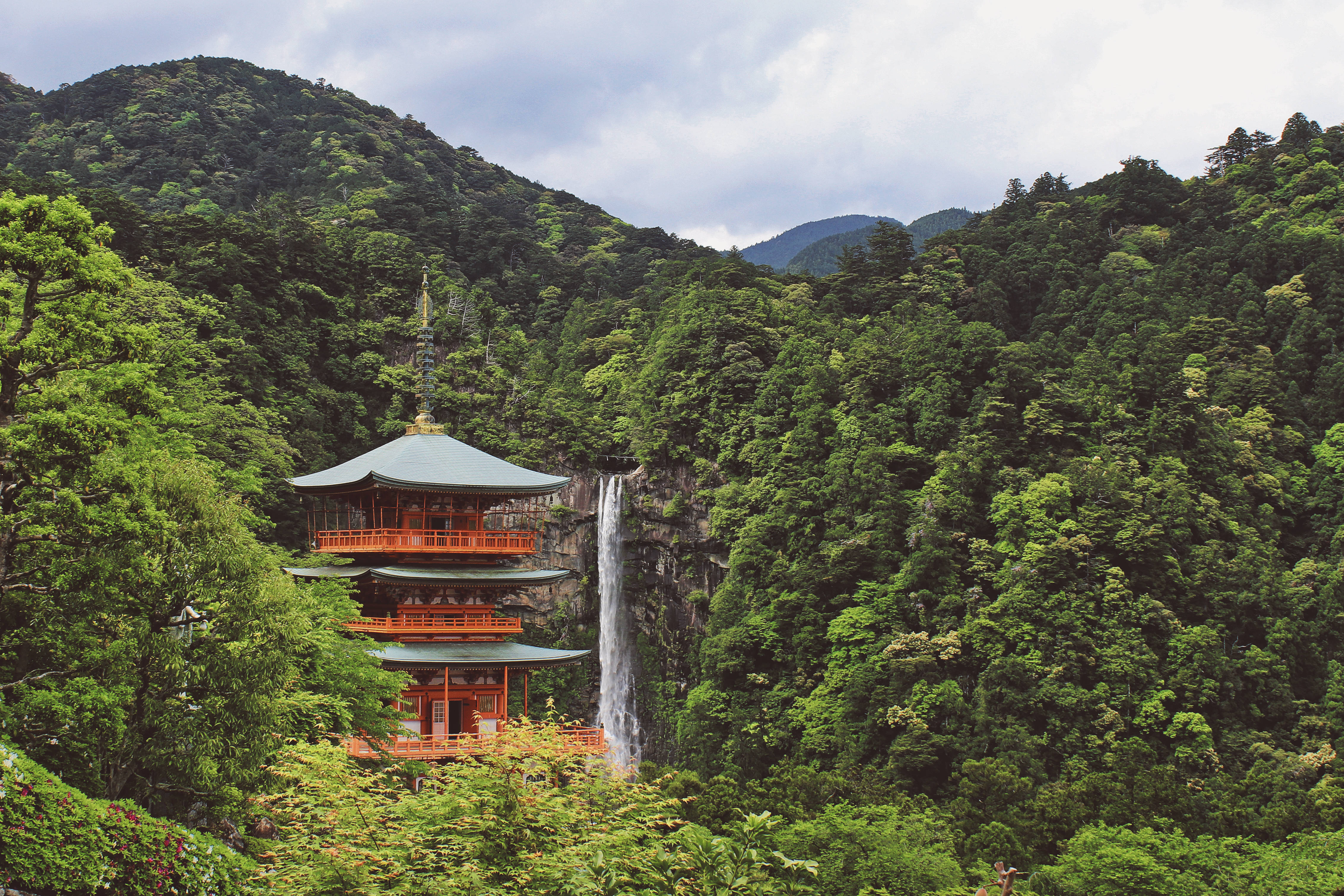日本 桌布 壁紙 — 一座傳統日本寺廟，紅色柱子和綠色屋頂，矗立在層疊瀑布旁，周圍是鬱鬱蔥蔥的綠色山巒。