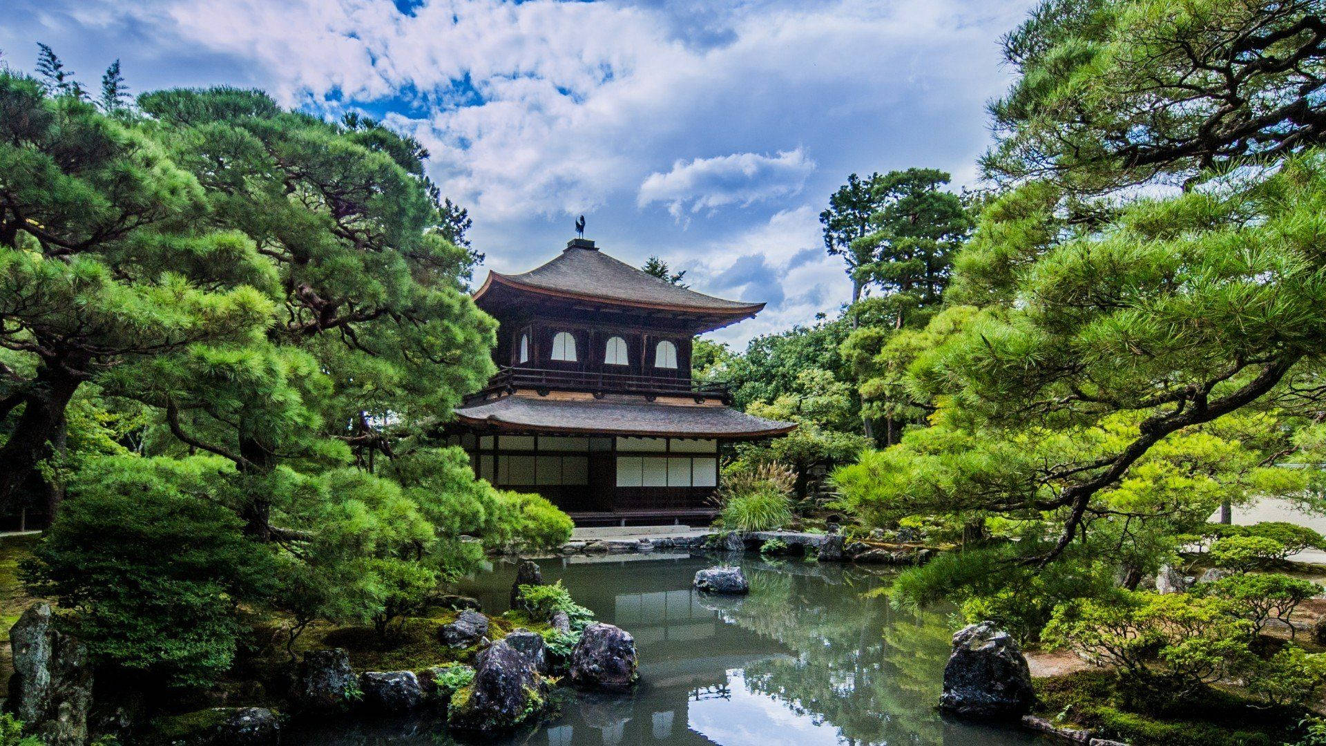 京都 桌布 壁紙 — 一座傳統日本寺廟亭閣，坐落在茂密的青松之間，倒映在平靜的池塘中，上方是明亮的藍天。