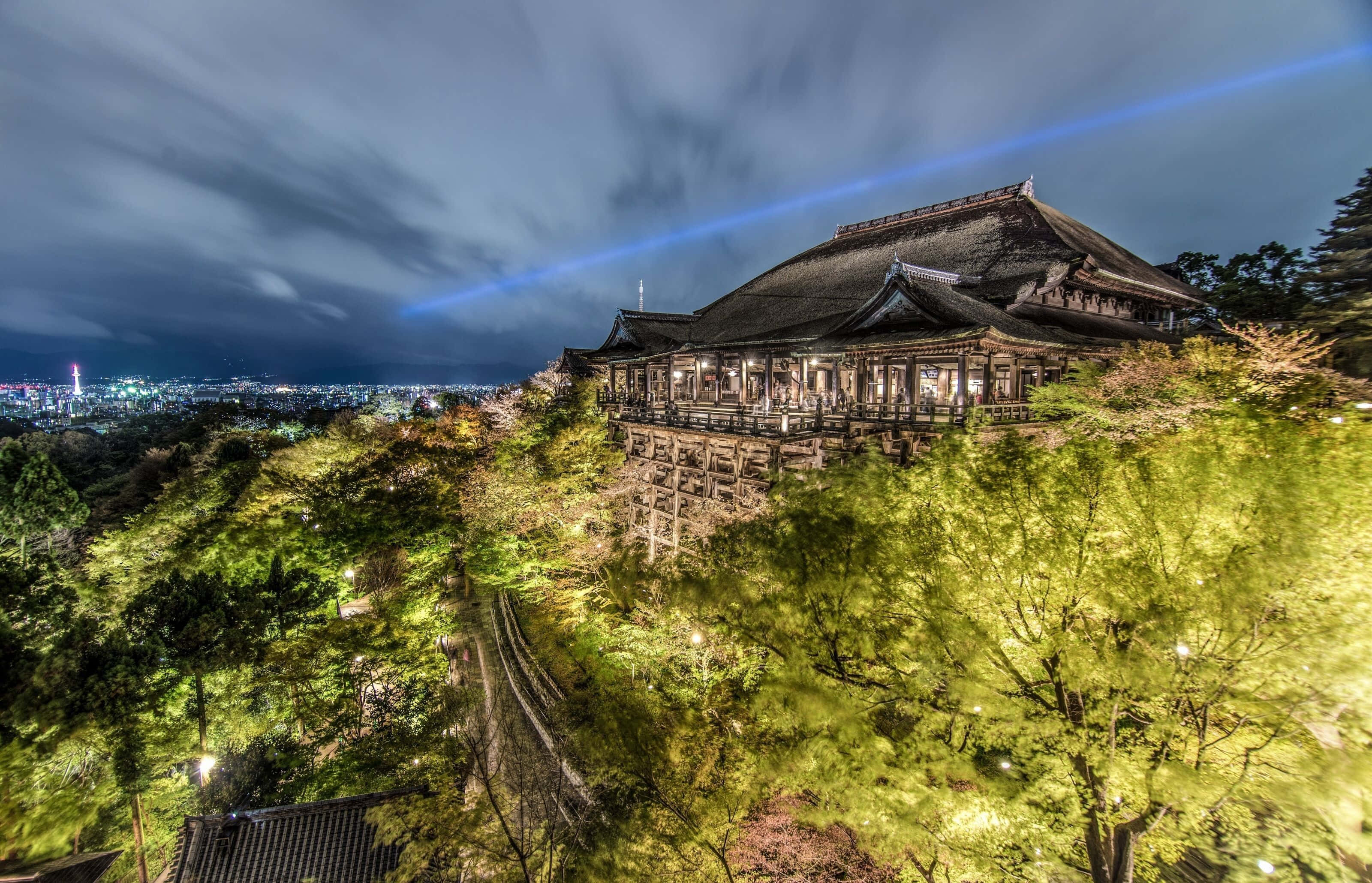 日本 桌布 壁紙 — 一座傳統日本寺廟坐落在山坡上，夜晚被溫暖的燈光照亮，映襯著佈滿雲層的黑暗天空和下方的城市燈光。
