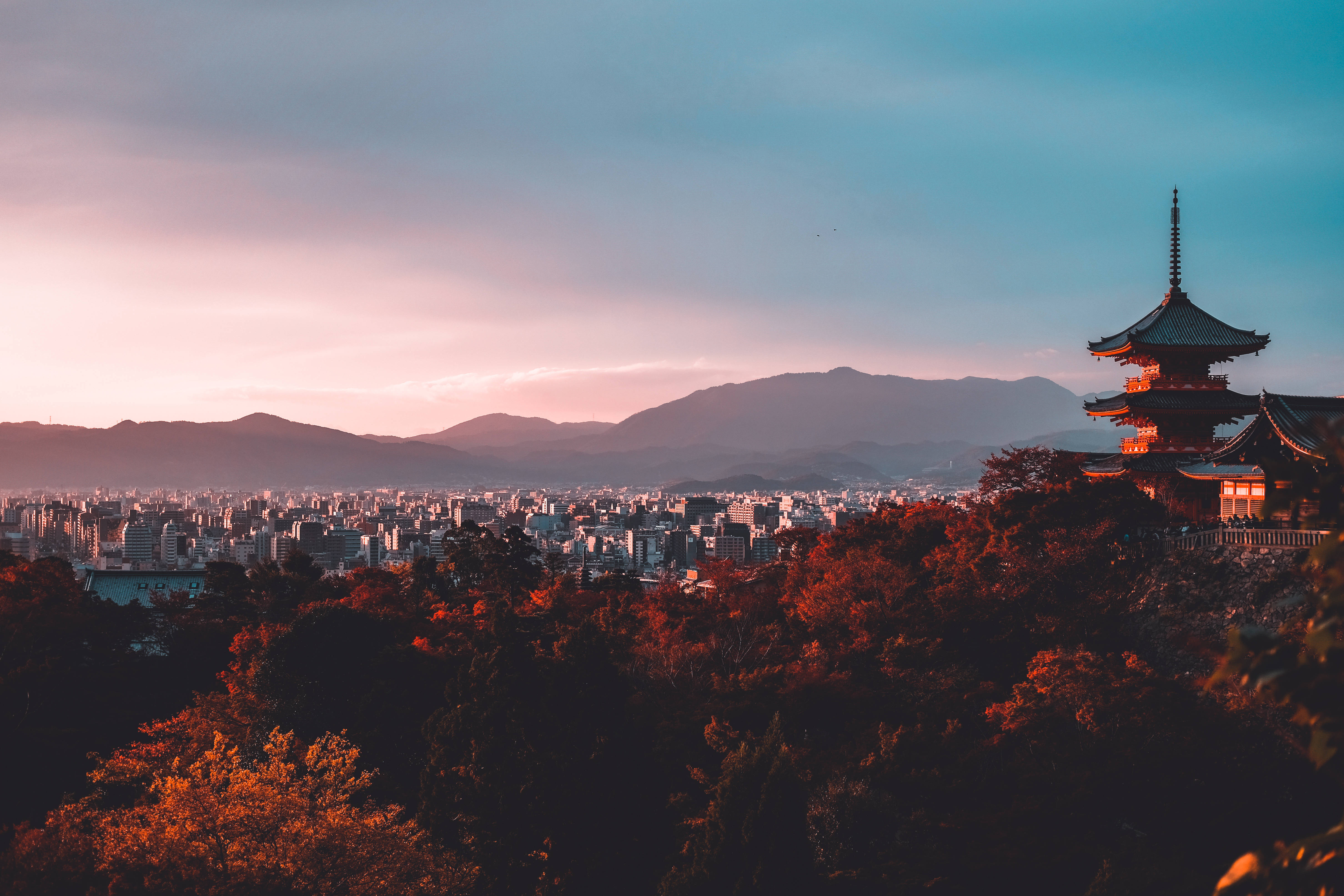 日本 桌布 壁紙 — 一座傳統日本寺廟坐落在秋樹之上，俯瞰著夕陽下廣闊的城市和遠處的山脈。