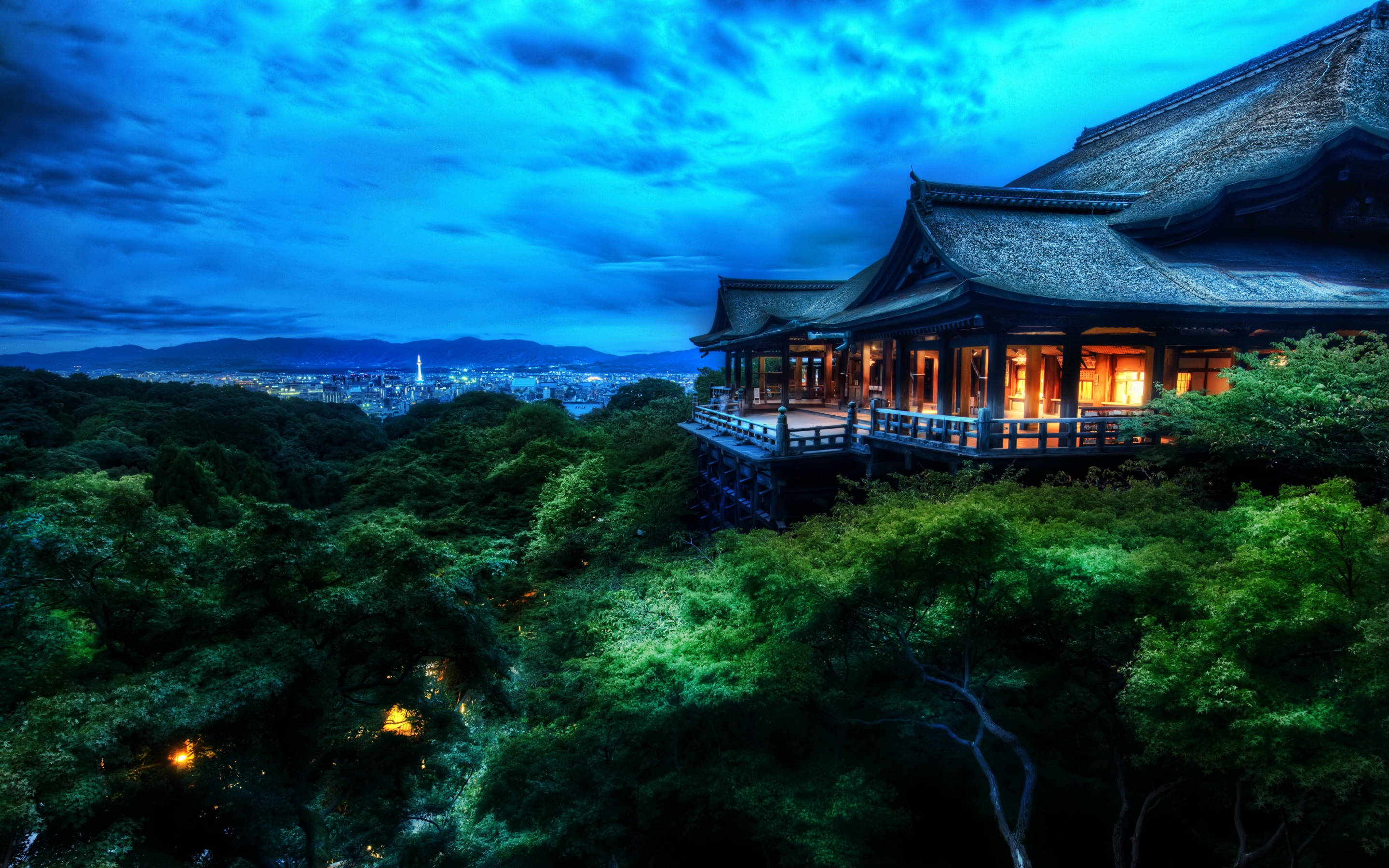 中式房屋壁紙 桌布 壁紙 — 一座傳統日本寺廟坐落在茂密的綠樹頂端，俯瞰著在戲劇性暮色天空下延伸的城市。