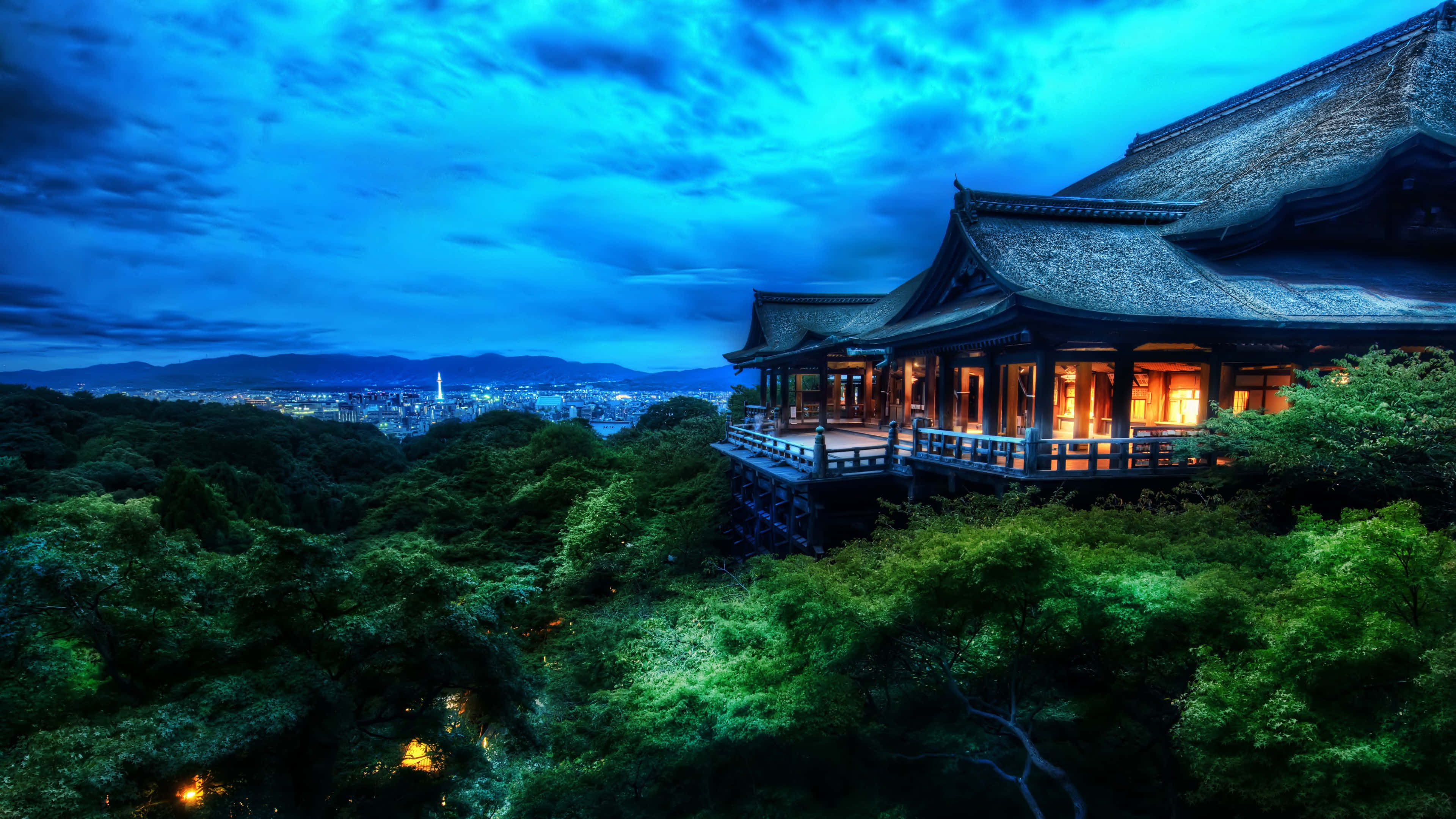 桌布 壁紙 — 一座傳統日本寺廟坐落在鬱鬱蔥蔥的山坡上，在深藍色的暮色天空和遠處的城市燈光中溫暖地發光。