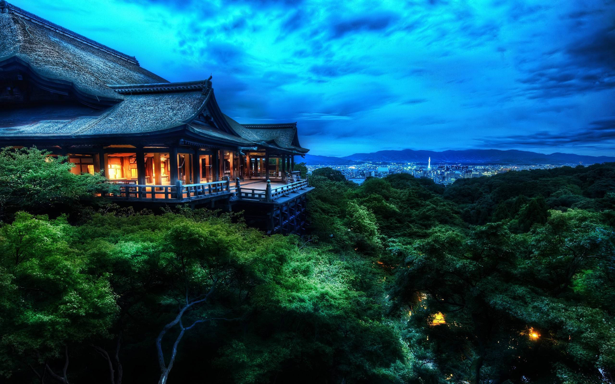 京都 桌布 壁紙 — 一座傳統木造寺廟坐落在林木茂密的山丘上，俯瞰著在戲劇性暮色天空下蔓延的城市。