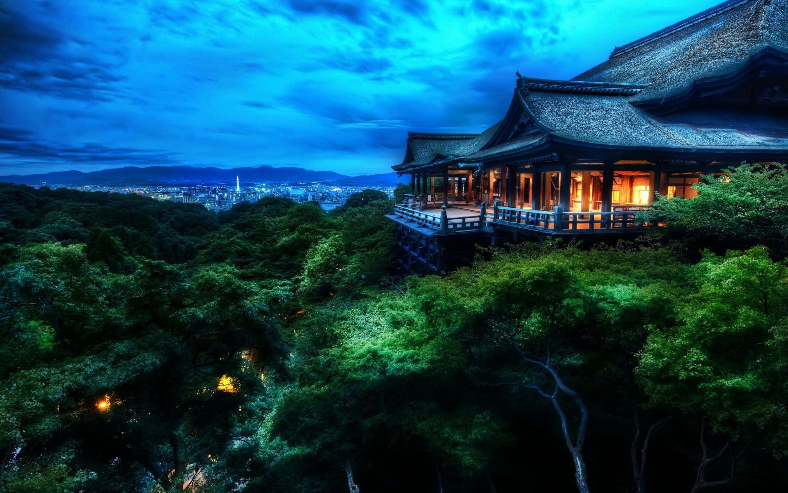 桌布 壁紙 — 一座傳統木造寺廟坐落在茂密的綠樹之上，在深藍色的暮色天空中溫暖地發光，俯瞰著遠處的城市景觀。