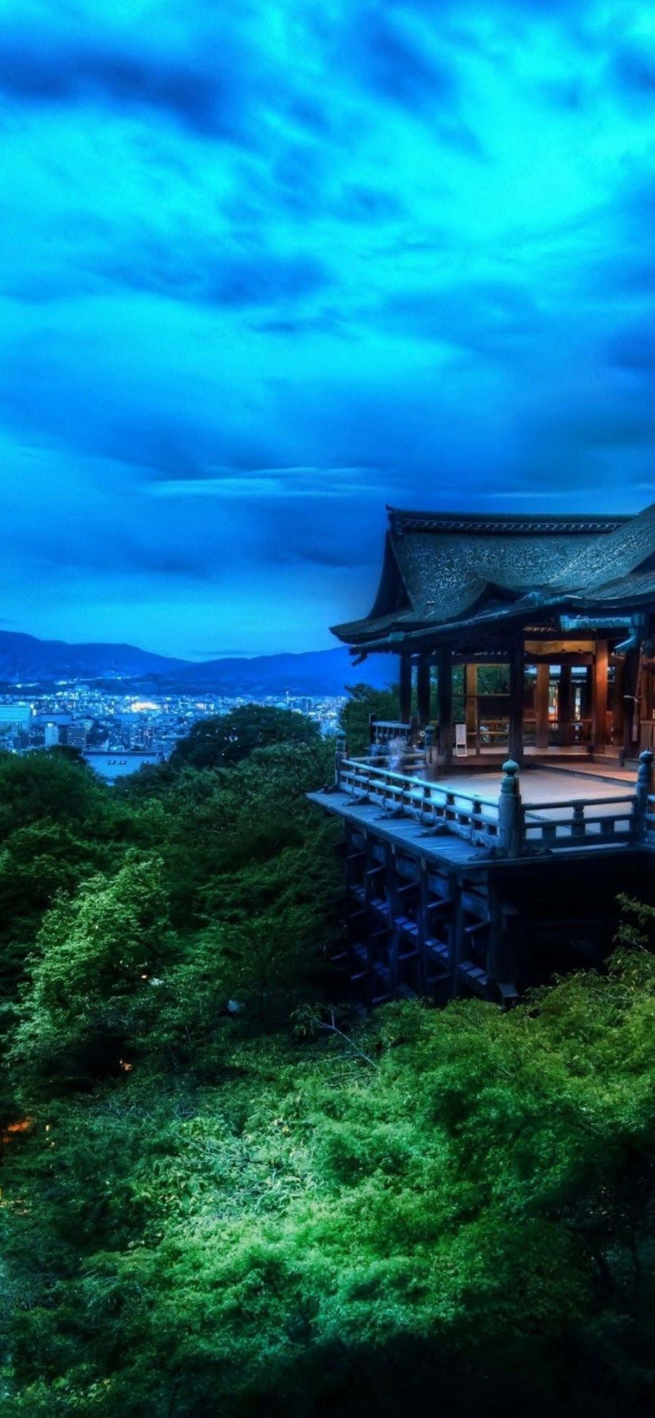 酷炫iPhone 桌布 壁紙 — 一座傳統木造寺廟坐落在蒼翠的山丘上，俯瞰著在戲劇性暮色天空下延伸的城市。