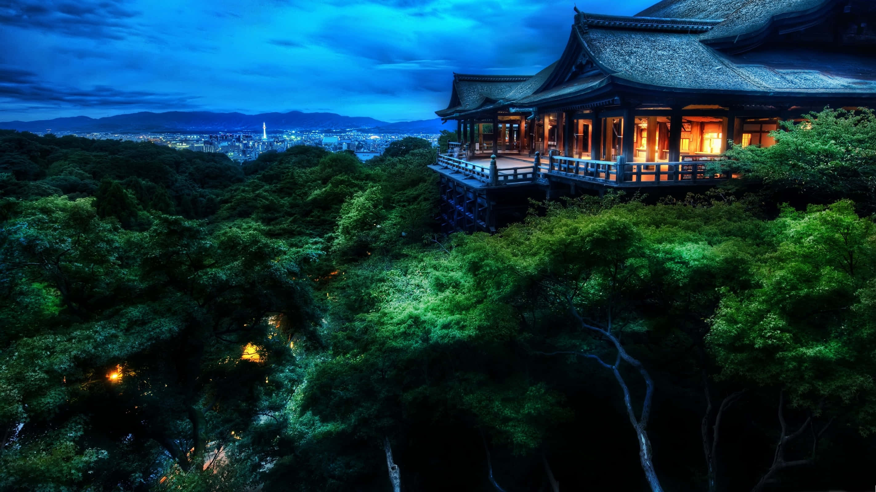 日本 桌布 壁紙 — 一座傳統木造寺廟坐落在鬱蔥的綠色山丘上，俯瞰著暮色中燈火通明的城市，窗戶在深藍色天空下閃爍著光芒。