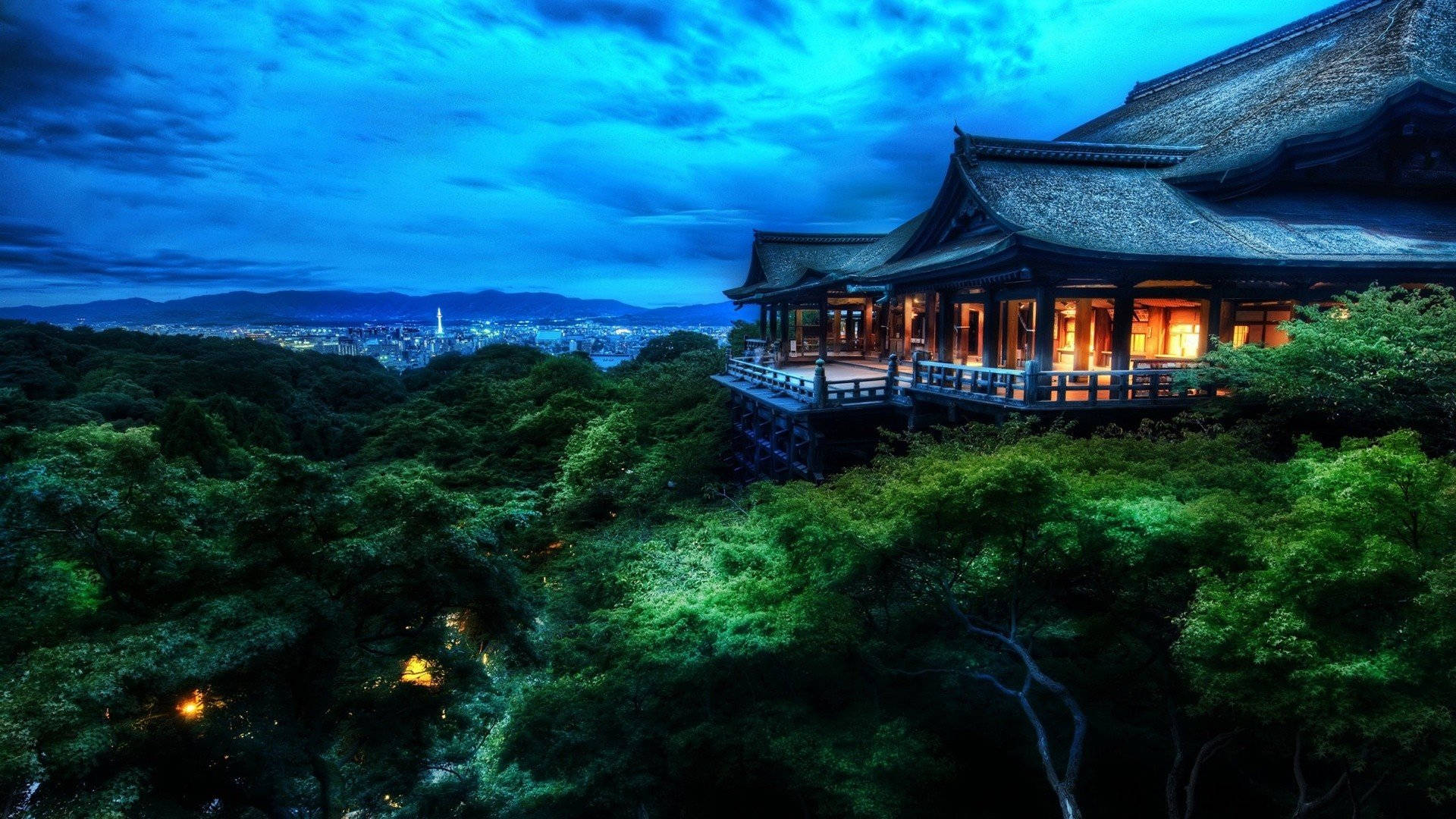日式自然壁紙 桌布 壁紙 — 一座傳統木造寺廟坐落在鬱鬱蔥蔥的山丘上，俯瞰著暮色中窗戶閃爍燈光的廣闊城市，背景是深藍色的天空。