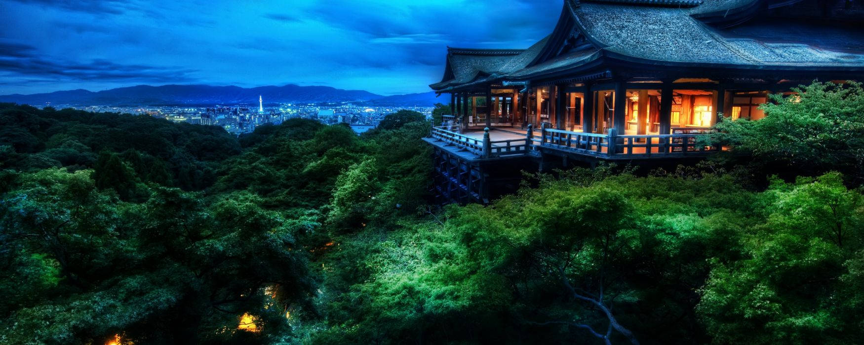 寬螢幕動態圖片 桌布 壁紙 — 一座傳統木造寺廟坐落在鬱鬱蔥蔥的山丘上，俯瞰著暮色中燈火通明的城市，窗戶閃爍，天空深藍。