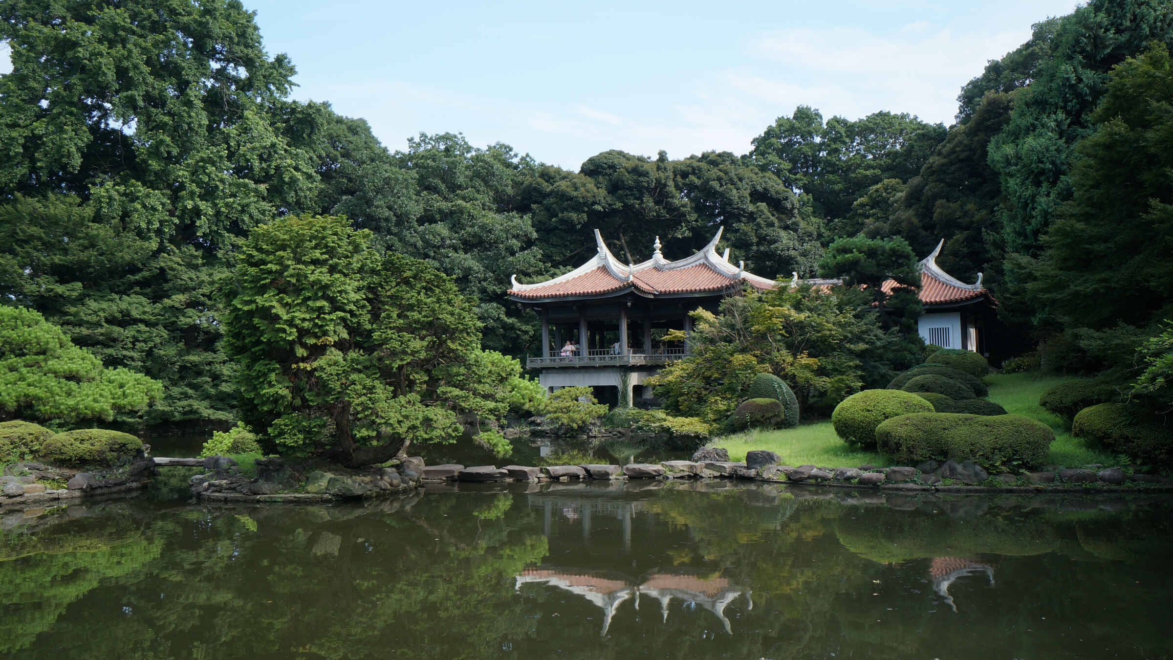 Fukuoka 桌布 壁紙 — 一座傳統東亞亭閣，擁有彎曲的屋頂，寧靜地坐落在平靜的池塘旁，倒映著茂密的綠樹和藍天。