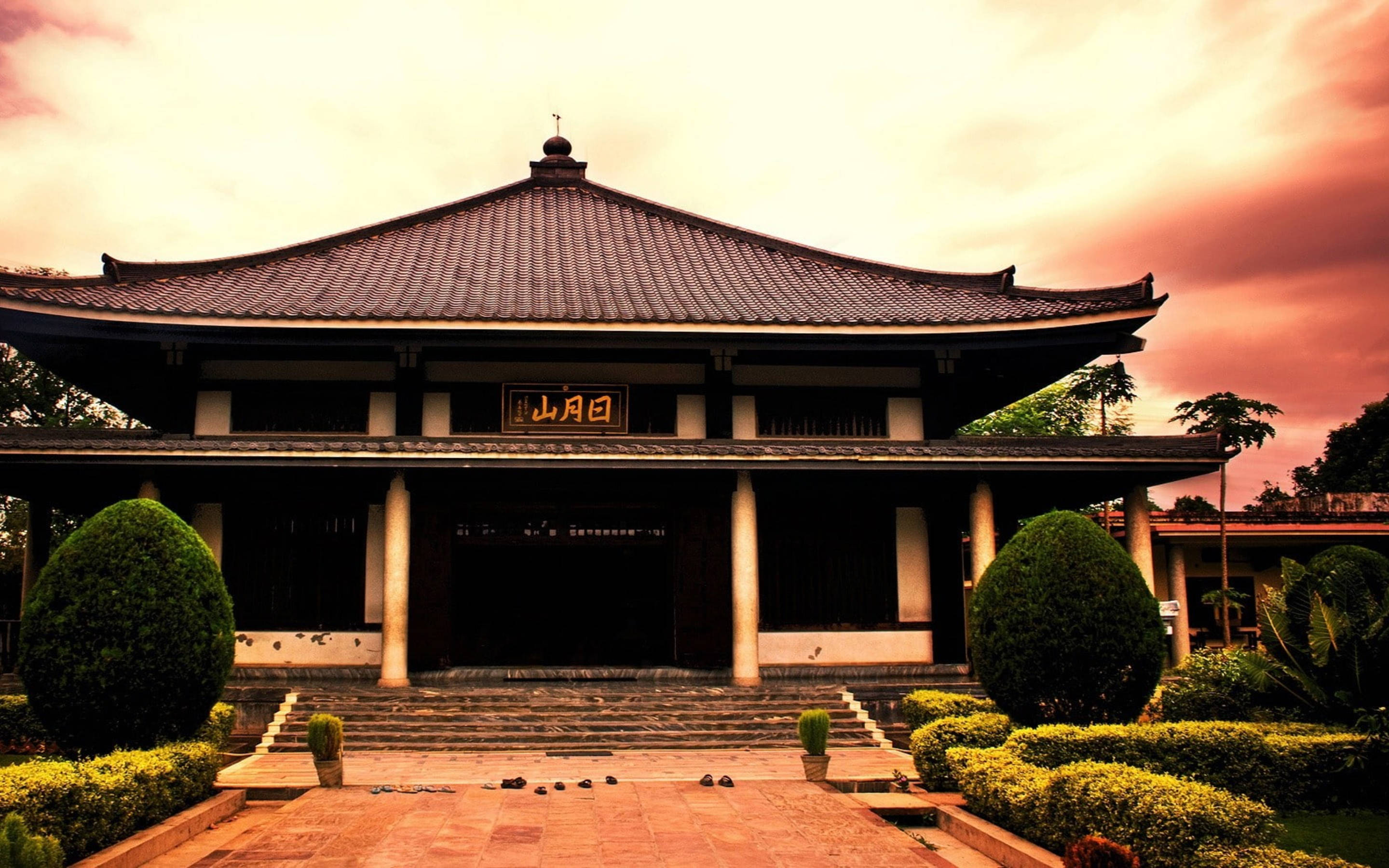 中式房屋壁紙 桌布 壁紙 — 一座傳統東亞寺廟，擁有瓦頂和石階，在戲劇性的日落天空下，由修剪整齊的灌木叢環繞。