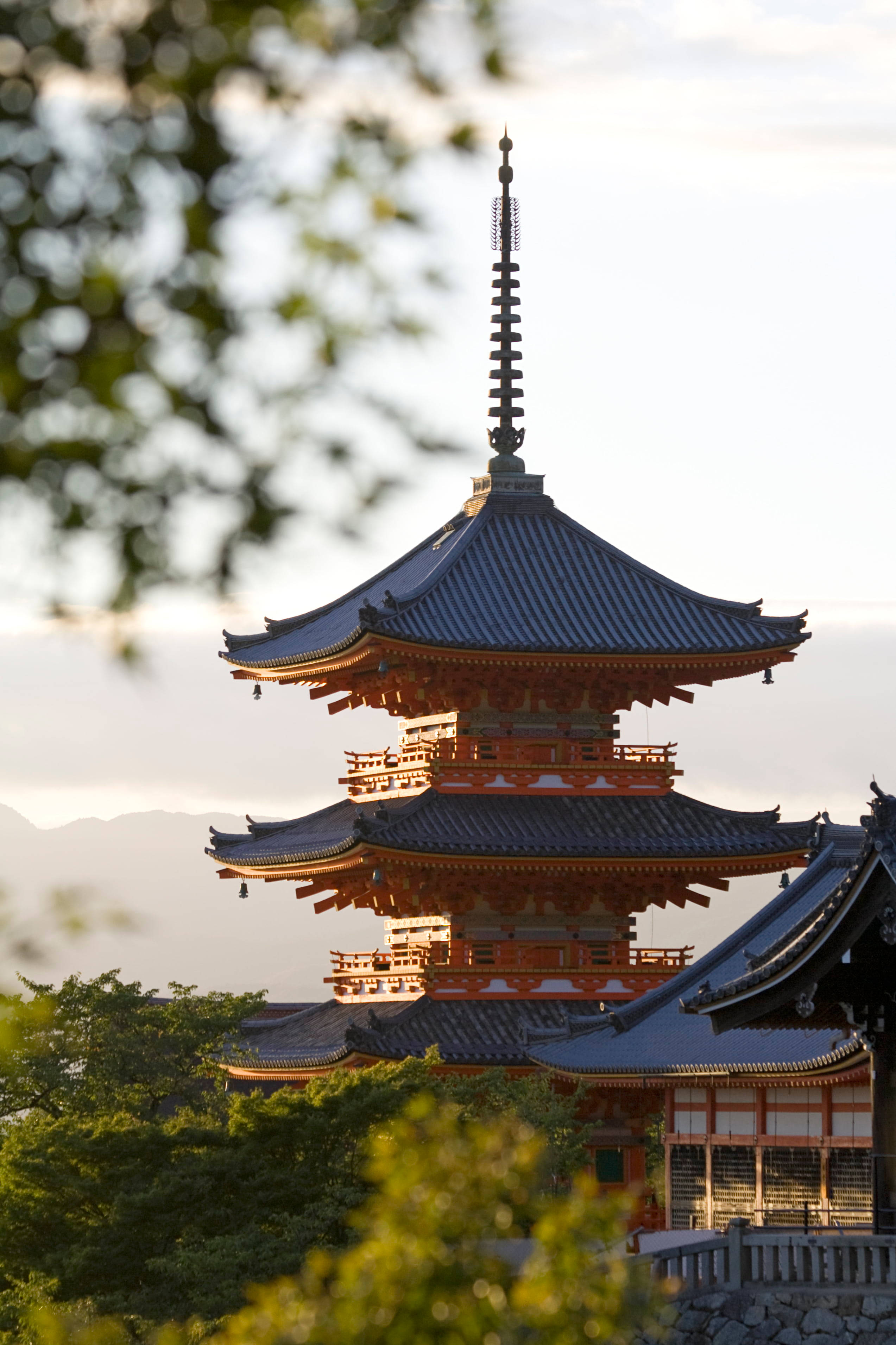 手機 桌布 壁紙 — 一座傳統的日本寶塔，層疊的屋頂和紅色木樑，在柔和的天空襯托下，被模糊的綠色樹葉框住，寧靜地矗立。