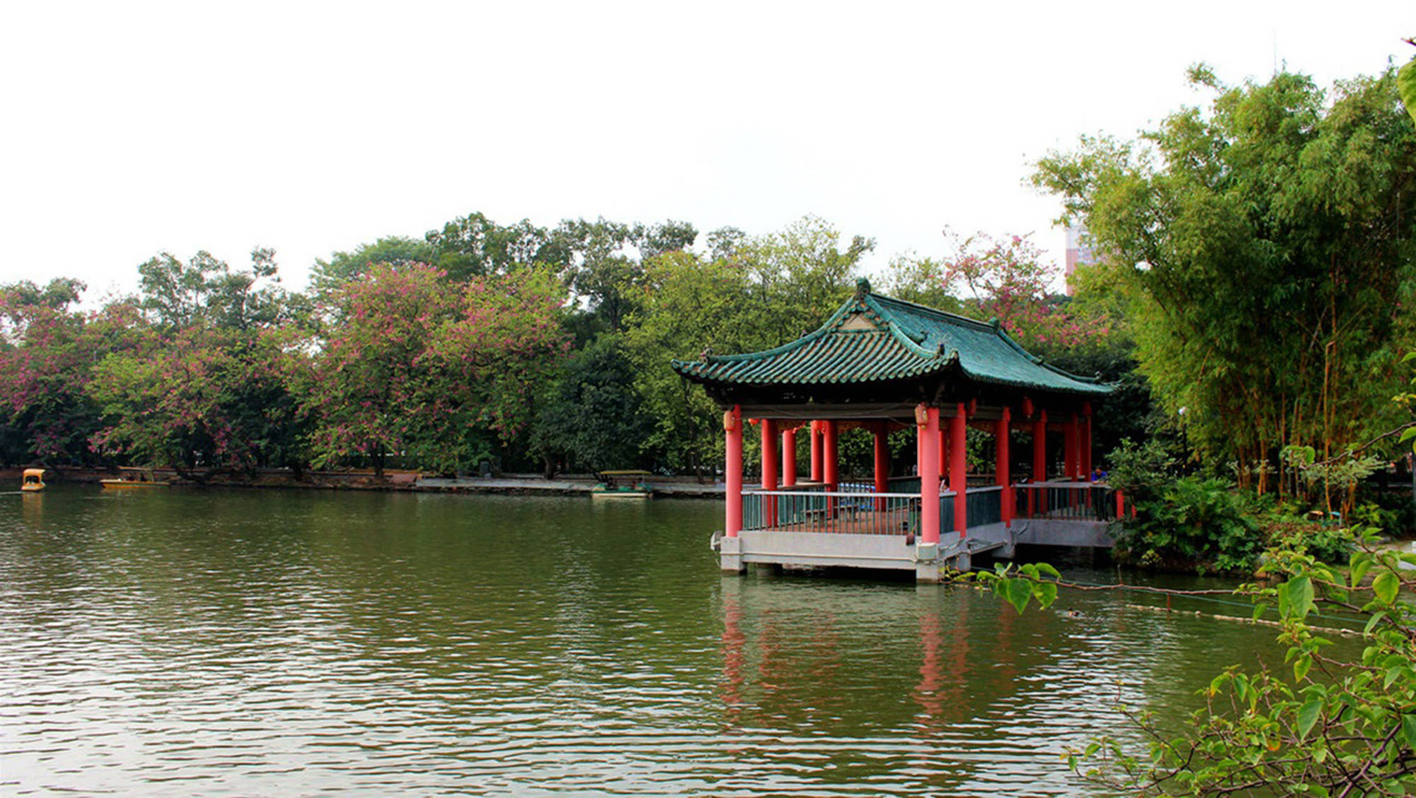 Guangzhou 桌布 壁紙 — 一座傳統的紅綠相間的中國亭子靜靜地漂浮在平靜的湖面上，周圍環繞著茂密的樹木和開花的灌木。