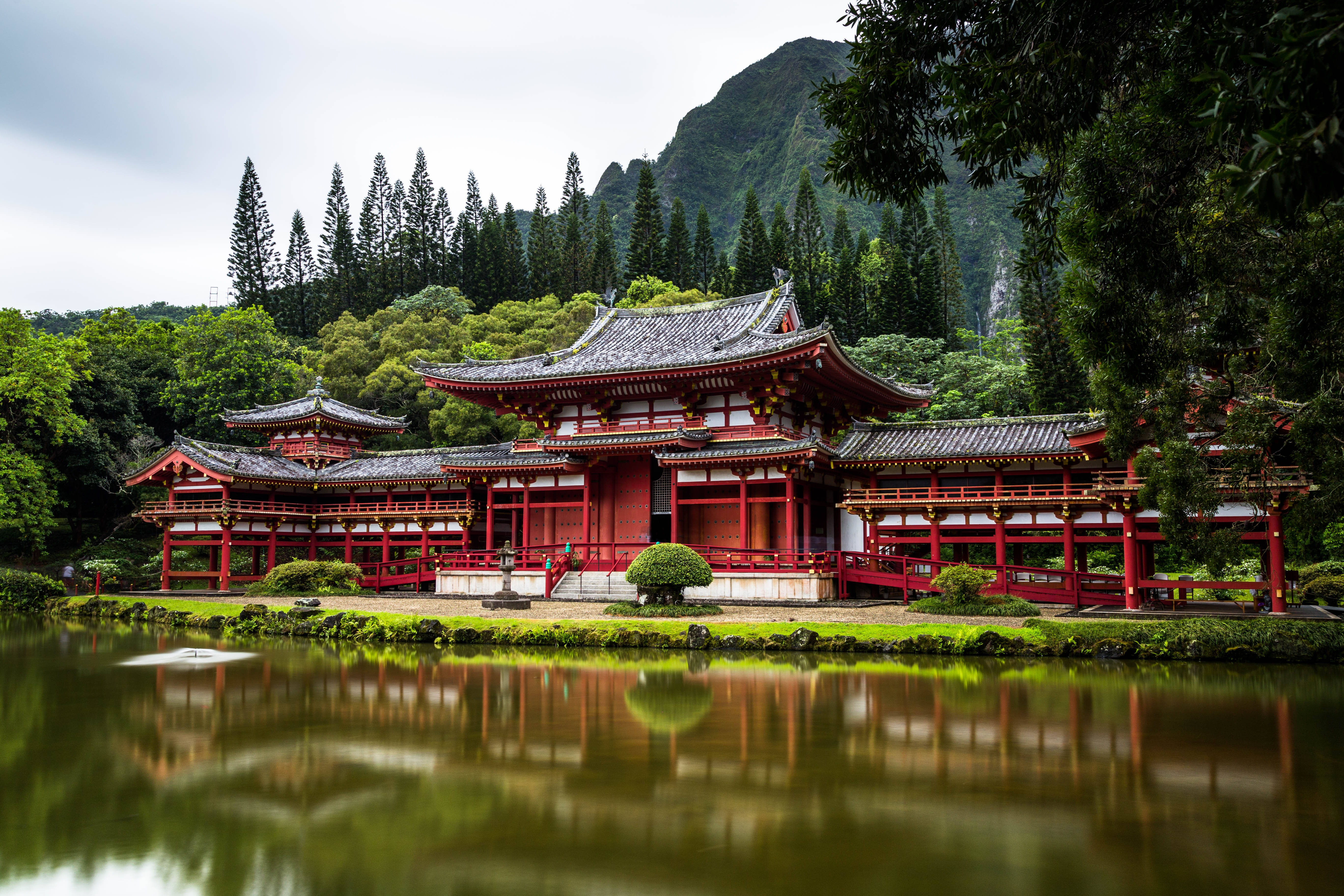 Japanese Aesthetic 桌布 壁紙 — 一座傳統的紅色日本寺廟，擁有彎曲的屋頂，倒映在平靜的水面上，周圍環繞著茂密的綠樹和山脈。