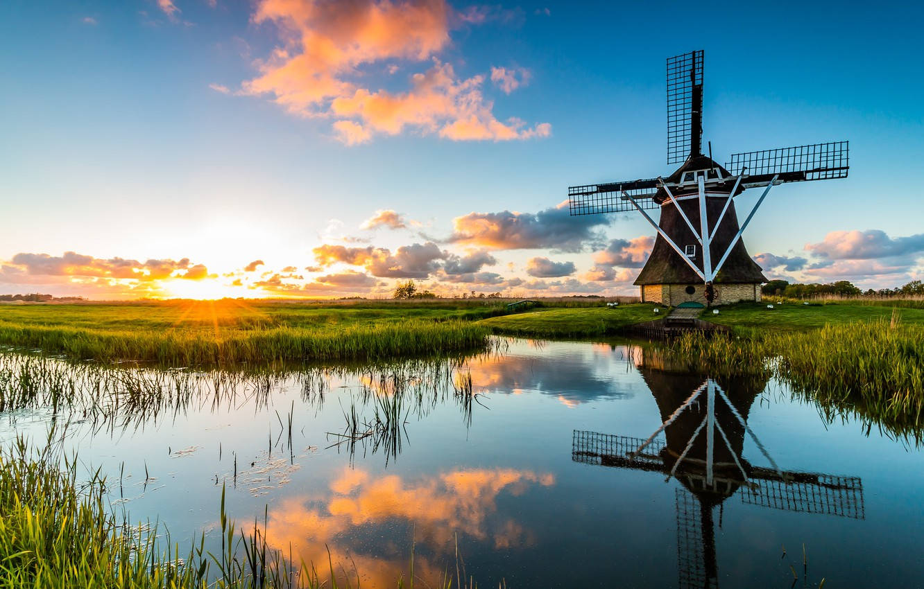 Netherlands 桌布 壁紙 — 一座傳統荷蘭風車在日落時高高聳立，其倒影完美地映照在平靜的水道上，周圍環繞著茂密的綠色蘆葦。