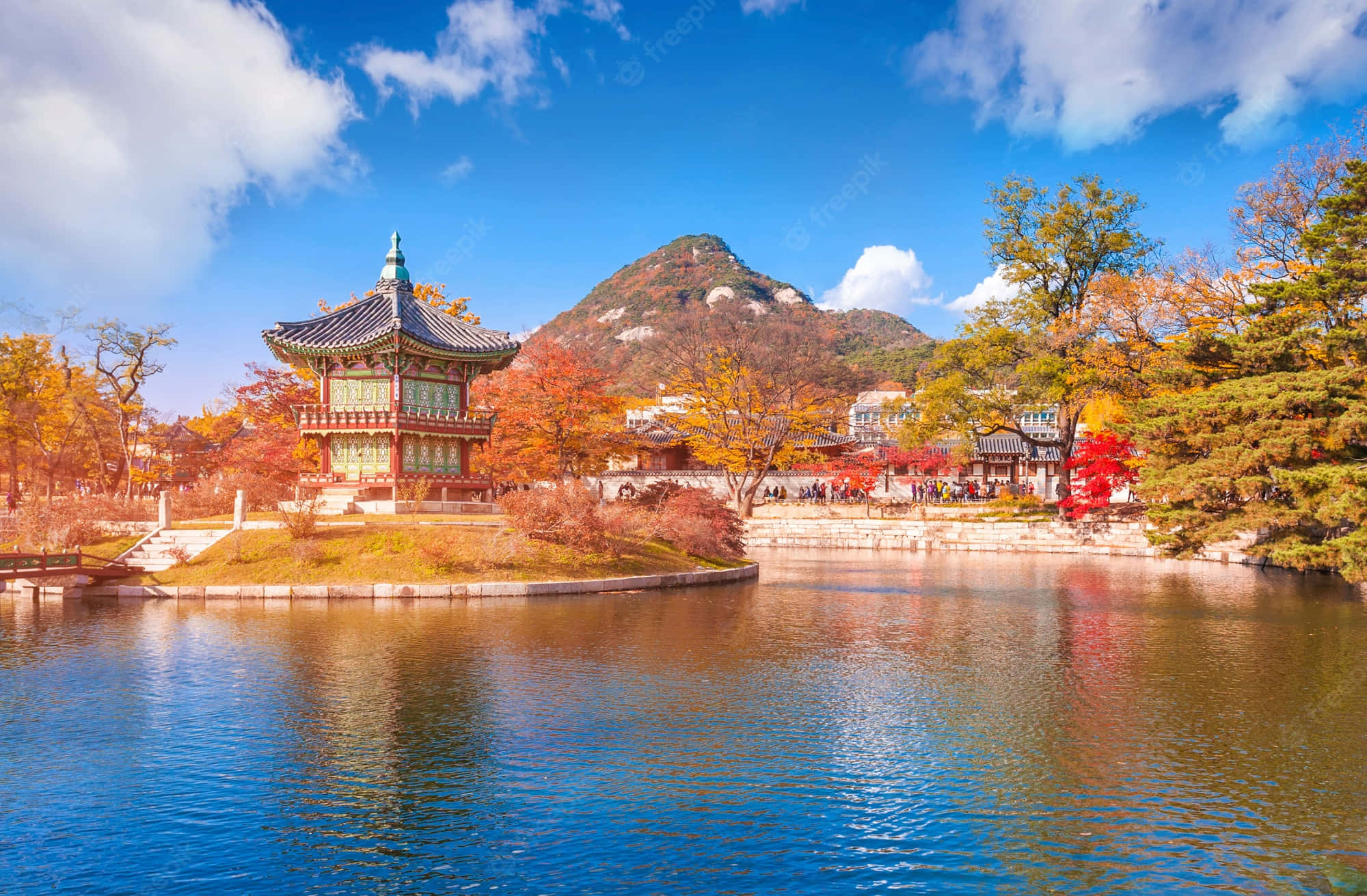 韓國 桌布 壁紙 — 一座傳統韓國亭子坐落在池塘上，周圍環繞著秋樹，樹葉呈現鮮豔的紅色、橙色和黃色，在明亮的藍天下。