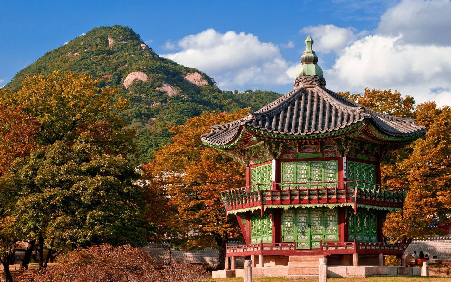 Tree 桌布 壁紙 — 一座傳統韓國亭閣，帶有綠色和紅色點綴，矗立在秋樹之間，背景是森林覆蓋的山脈，藍天白雲。