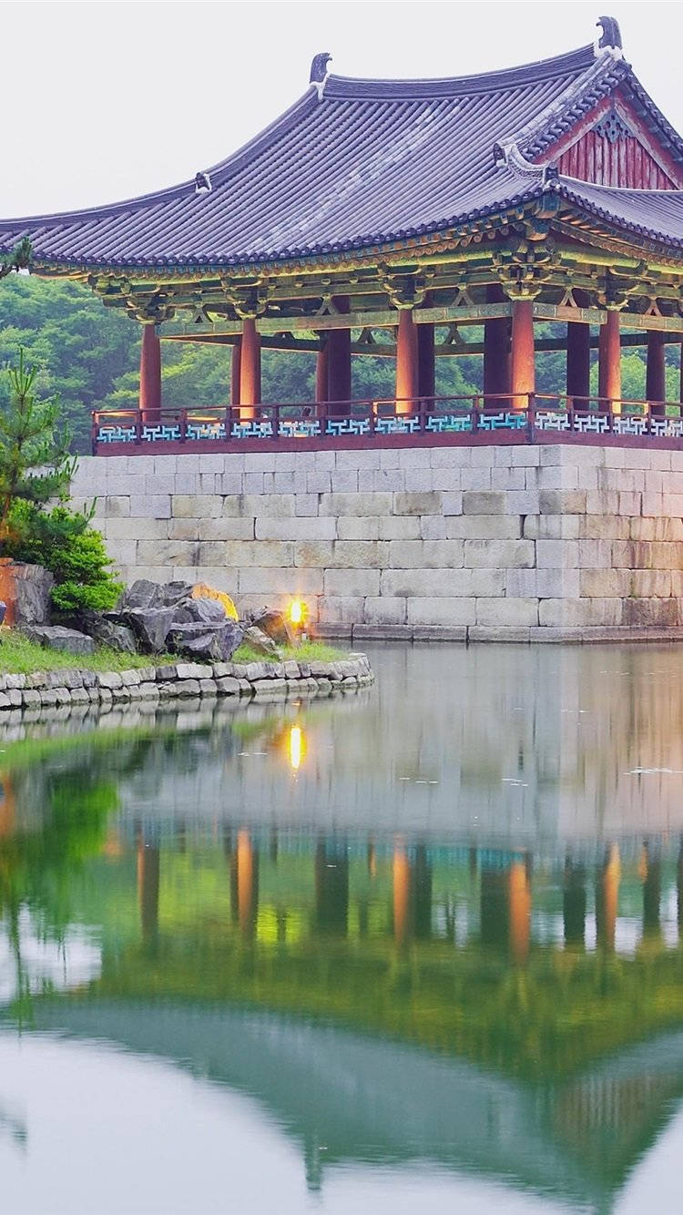 韓國 桌布 壁紙 — 一座傳統韓國亭閣，擁有彎曲的屋頂和紅色柱子，矗立在平靜的池塘旁，倒映出其寧靜的輪廓。