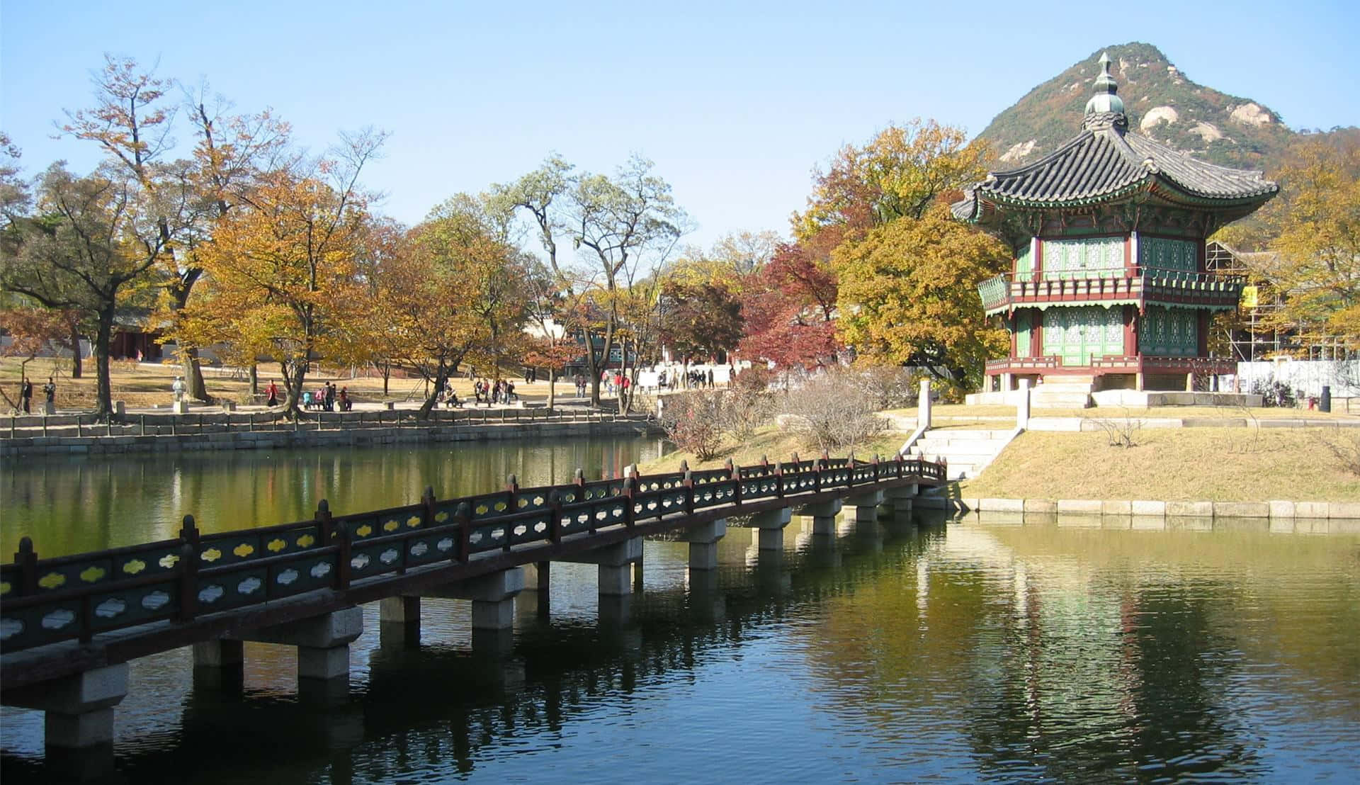 韓國 桌布 壁紙 — 一座傳統韓國亭閣矗立在平靜的池塘旁，有一座木橋，周圍環繞著秋天的樹木和遠處的山巒，在清澈的藍天下。