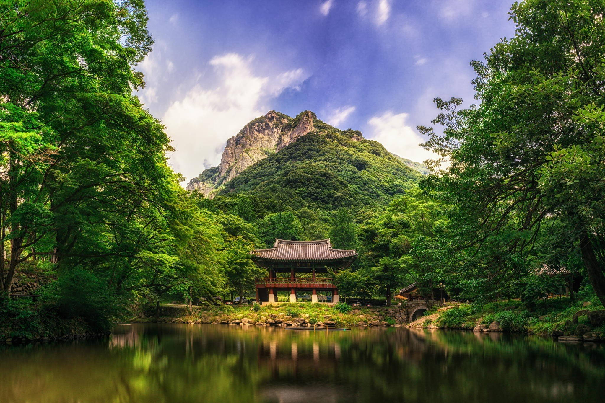山脈 桌布 壁紙 — 一座傳統韓國亭閣靜靜地坐落在平靜的池塘邊，四周環繞著茂密的綠樹和巍峨的山脈，上方是湛藍的天空。