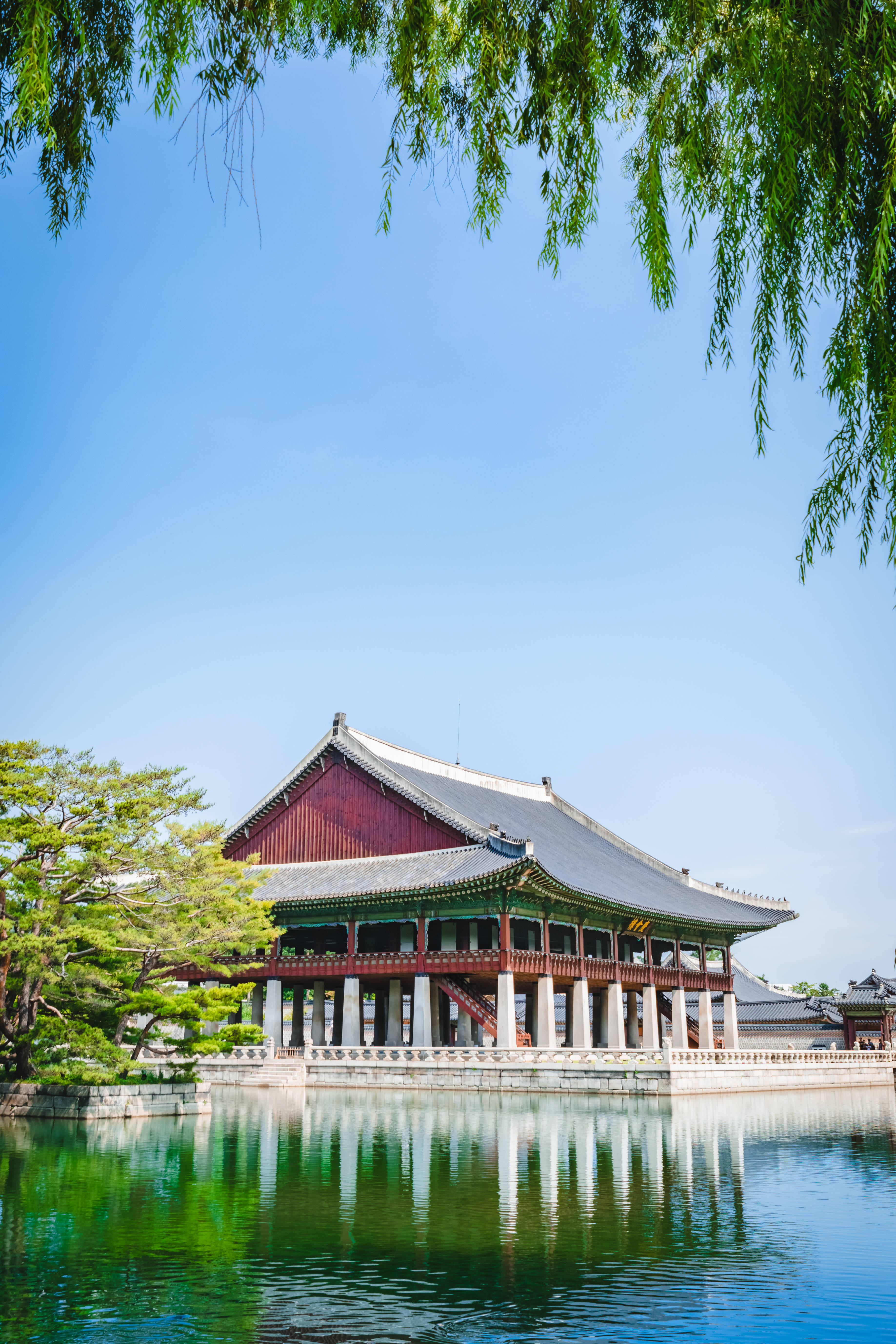南韓 桌布 壁紙 — 一座傳統韓國宮殿亭閣，紅色柱子灰色屋頂，在平靜的水面上倒映，藍天清澈，垂柳枝條環繞。