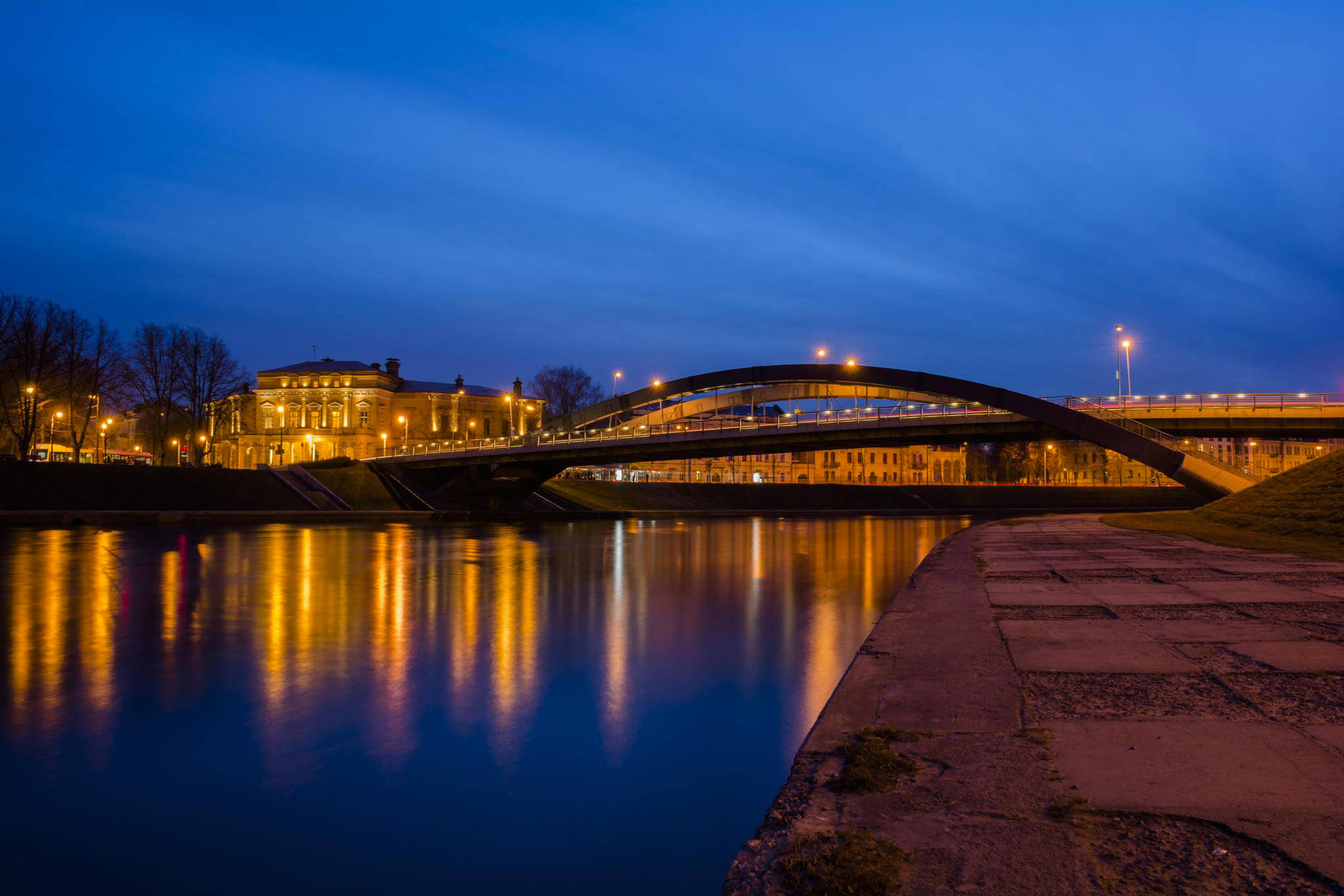 Vilnius 桌布 壁紙 — 一座優雅的拱橋在黃昏時分用溫暖的燈光照亮平靜的河流，映照著深藍色的天空。