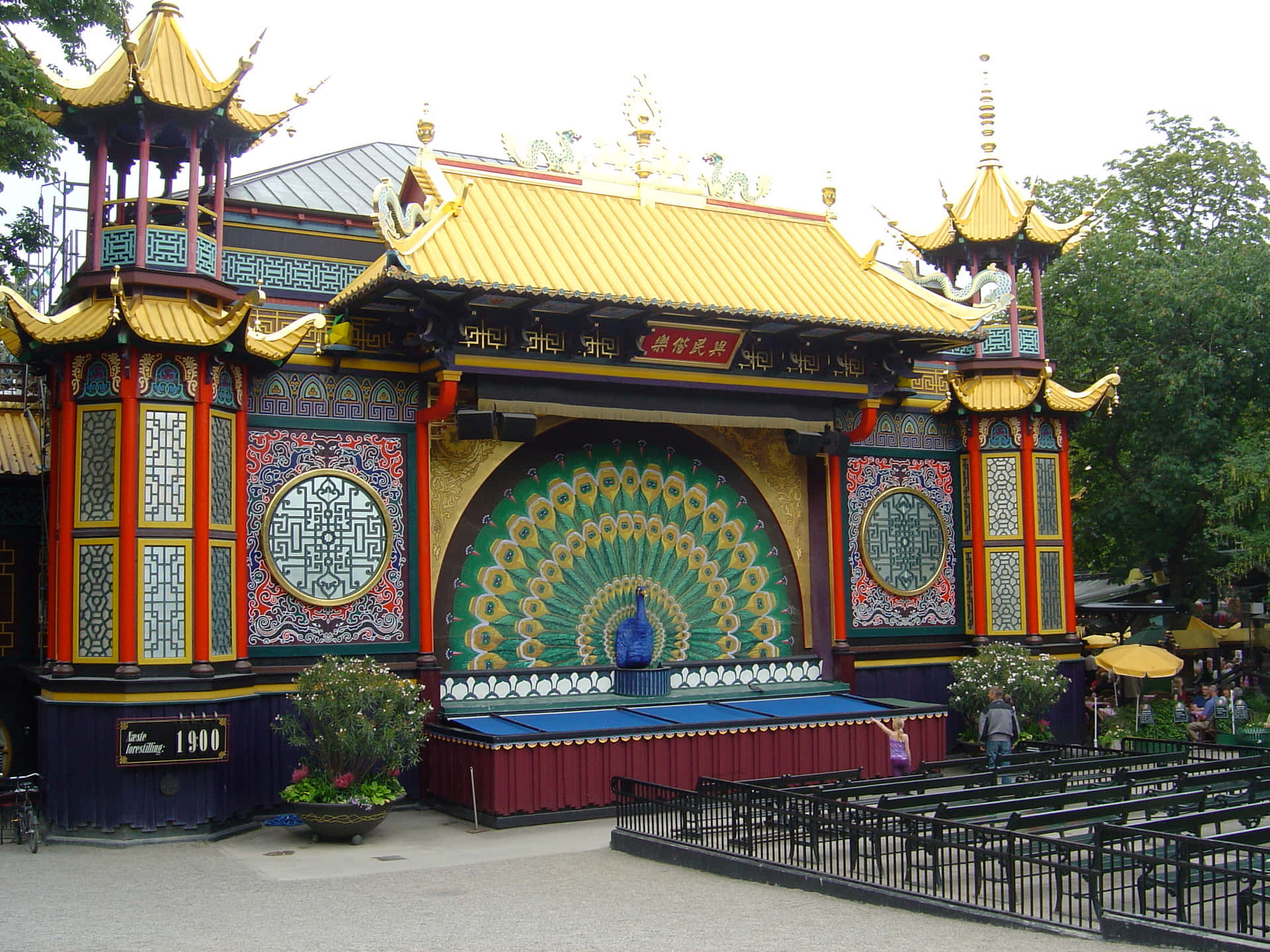 Theater 桌布 壁紙 — 一座充滿活力的中式亭閣，金色屋頂和大型孔雀壁畫，矗立在公園中，周圍有綠樹和遊客。