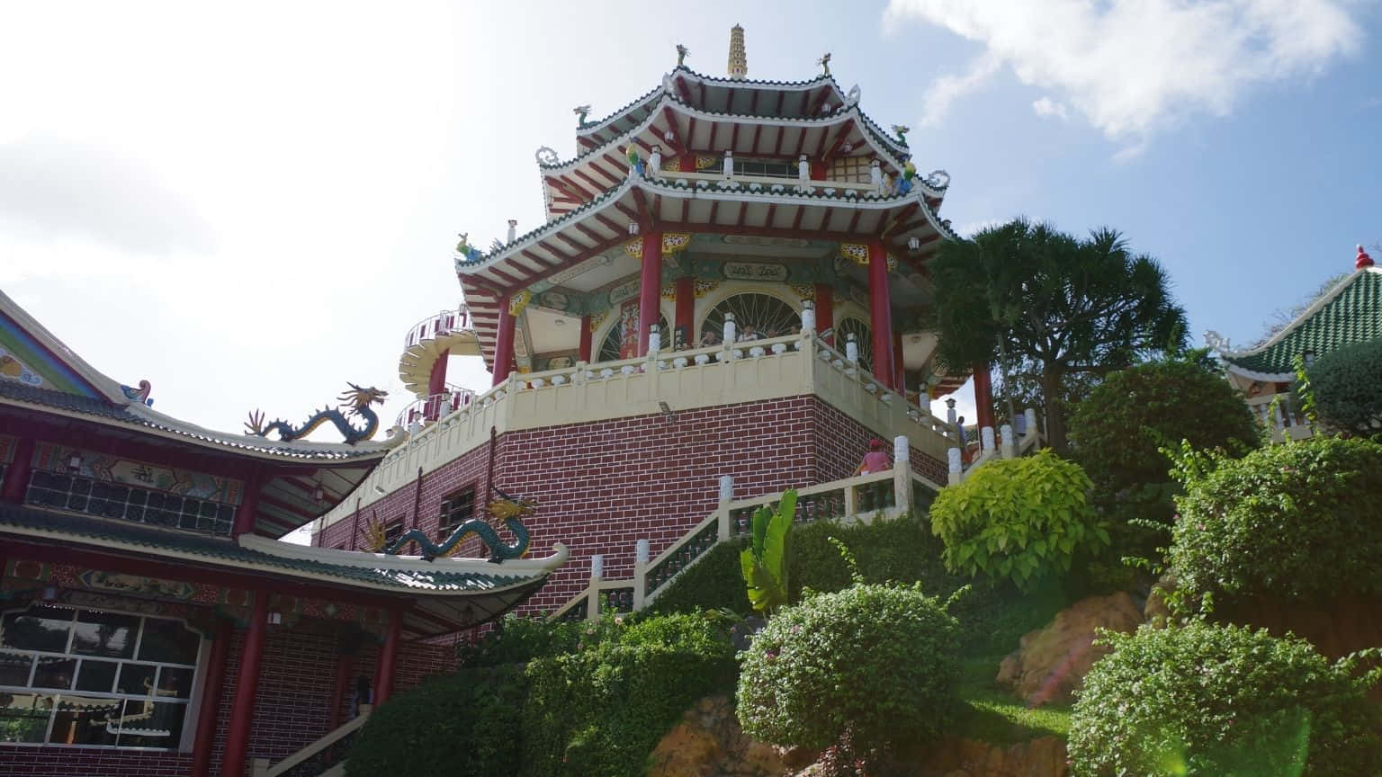 1536x864 桌布 壁紙 — 一座充滿活力的傳統中國寺廟，擁有紅色柱子和綠色屋頂，坐落在茂密的山坡花園頂部，在明亮的藍天下。