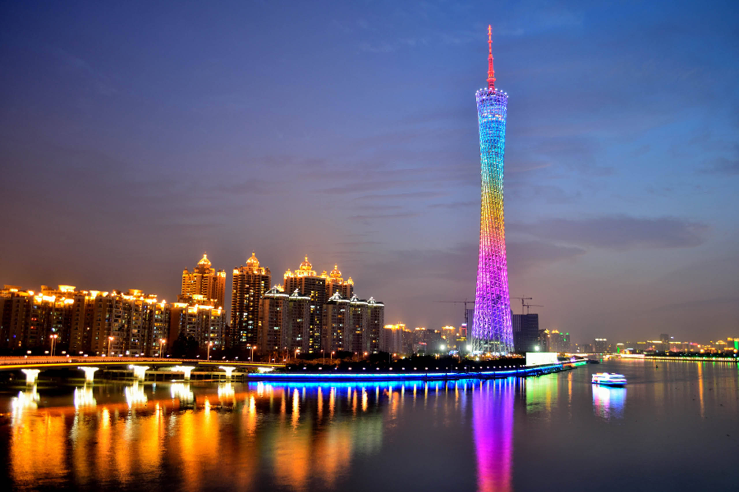 Guangzhou 桌布 壁紙 — 一座充滿活力的彩虹燈光塔在黃昏時分聳立於城市天際線之上，其燈光倒映在平靜的水面上，附近建築物閃閃發光。