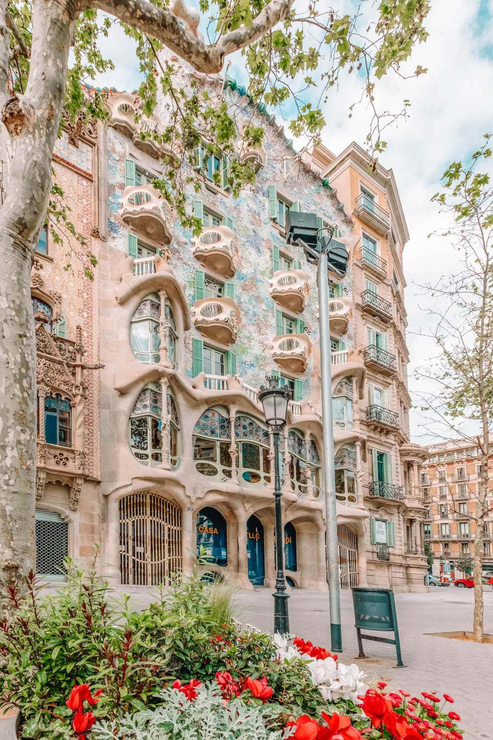 Barcelona 桌布 壁紙 — 一座充滿活力的新藝術風格建築，擁有有機曲線和馬賽克瓷磚，在部分多雲的天空下高高聳立，前景的綠樹成蔭和色彩繽紛的花壇襯托著它。