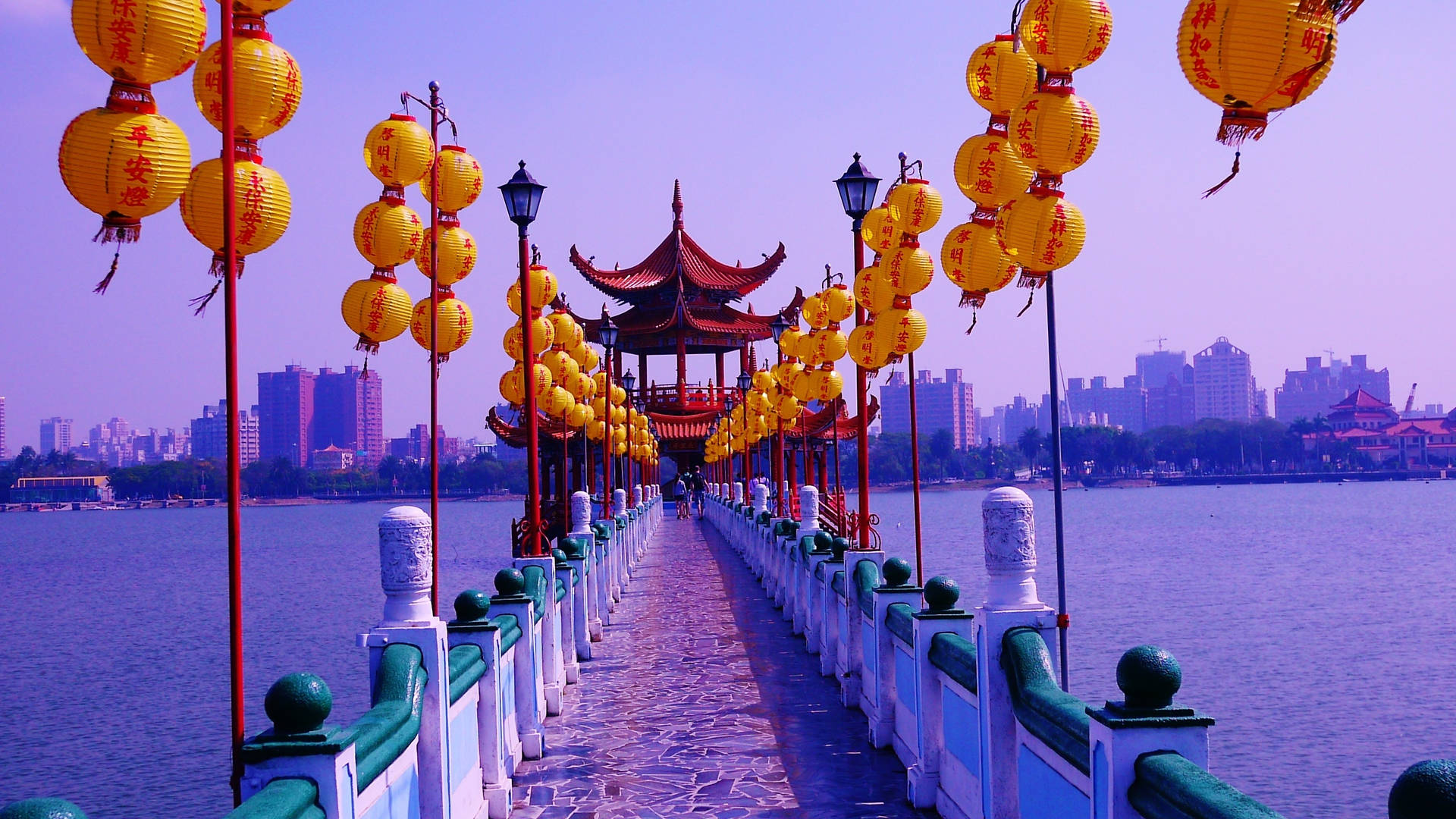 一座裝飾著黃色燈籠的橋樑橫跨水面，通往一座背景有城市建築的亭閣。