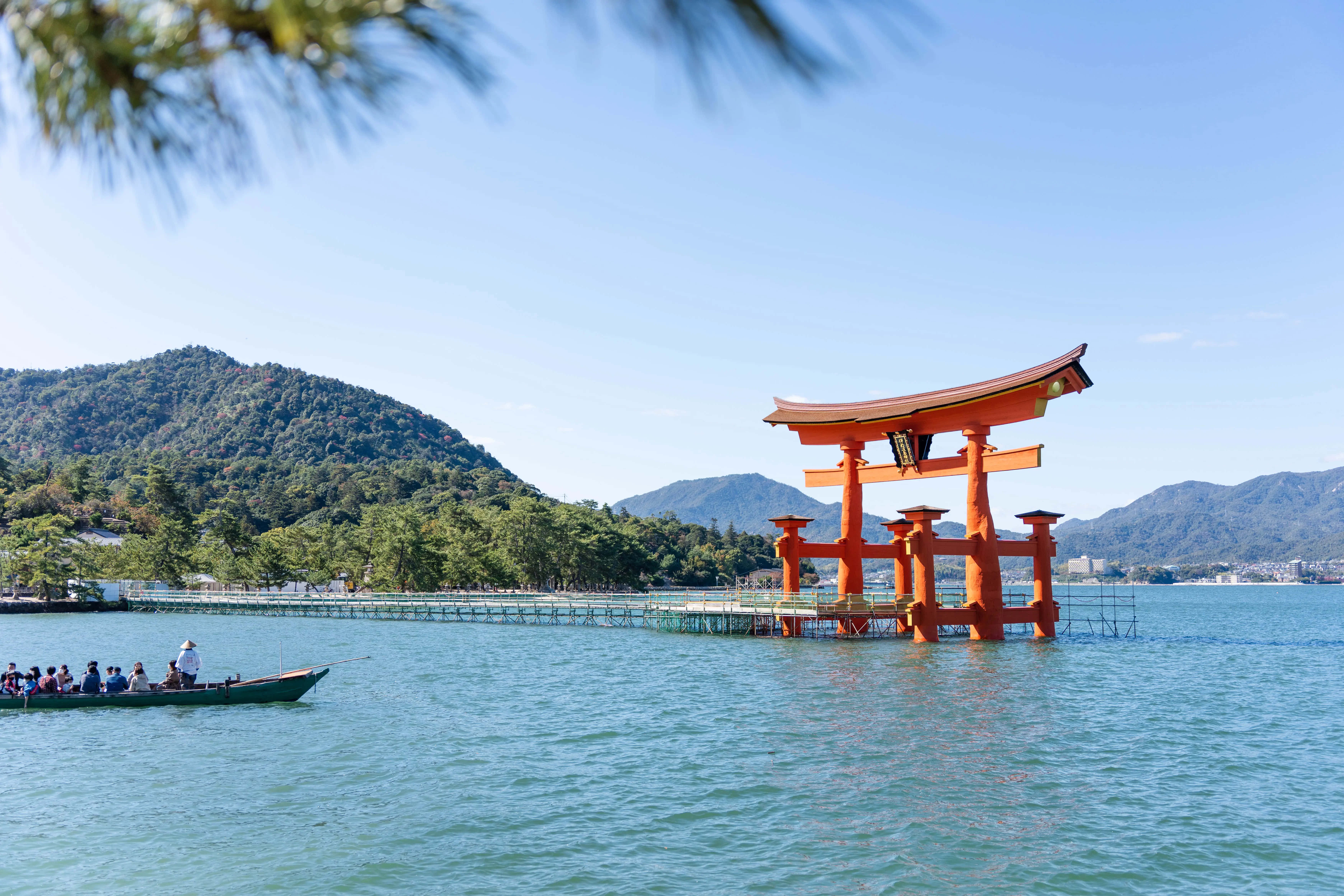 Travel And Tourism 桌布 壁紙 — 一座充滿活力的橙色鳥居雄偉地矗立在平靜的碧綠海水中，附近有一艘載著遊客的船隻，在晴朗的藍天下航行。